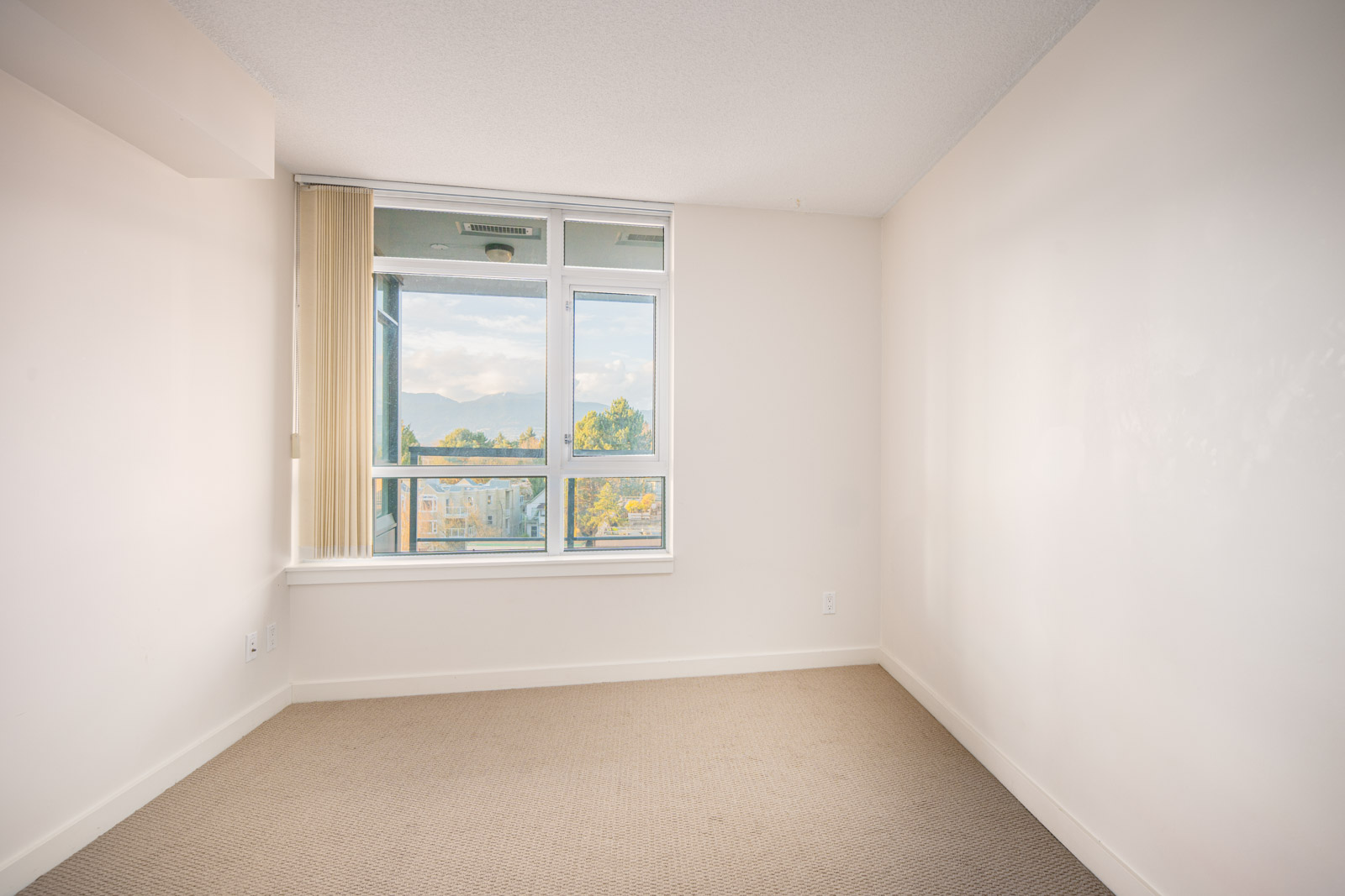 Empty room with beige carpet, white walls, and a large window with vertical blinds, showing a view of buildings and trees outside.