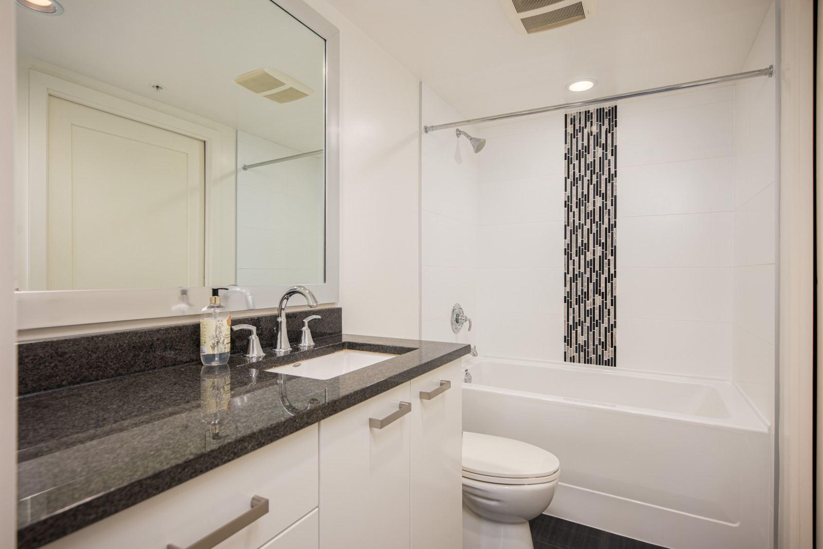 Modern bathroom with white walls, black countertop, sink, toilet, and bathtub featuring a vertical black and white tile accent in the shower area.