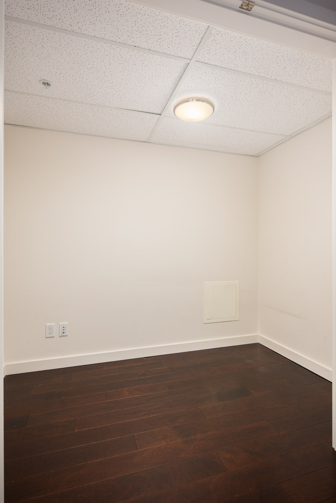 Small, empty room with white walls, dark wood flooring, a recessed ceiling light, and a square wall panel. Two electrical outlets are visible on the left wall.