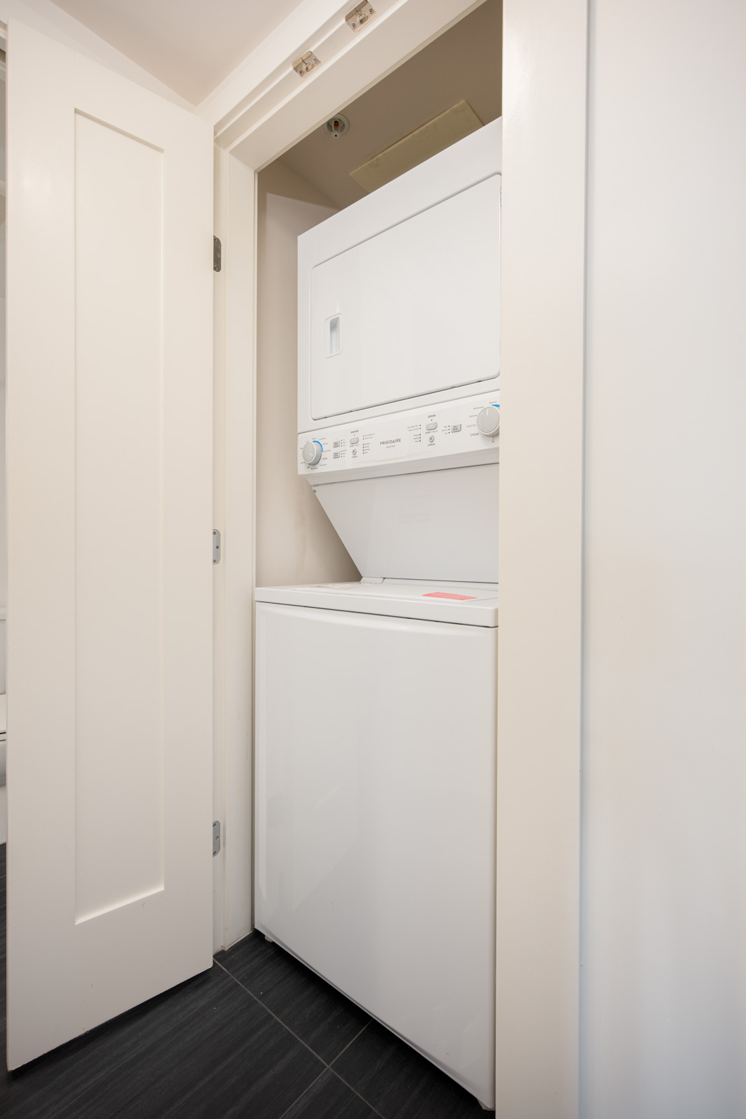 Stacked washer and dryer unit in a small laundry closet with white folding doors and black tile flooring.