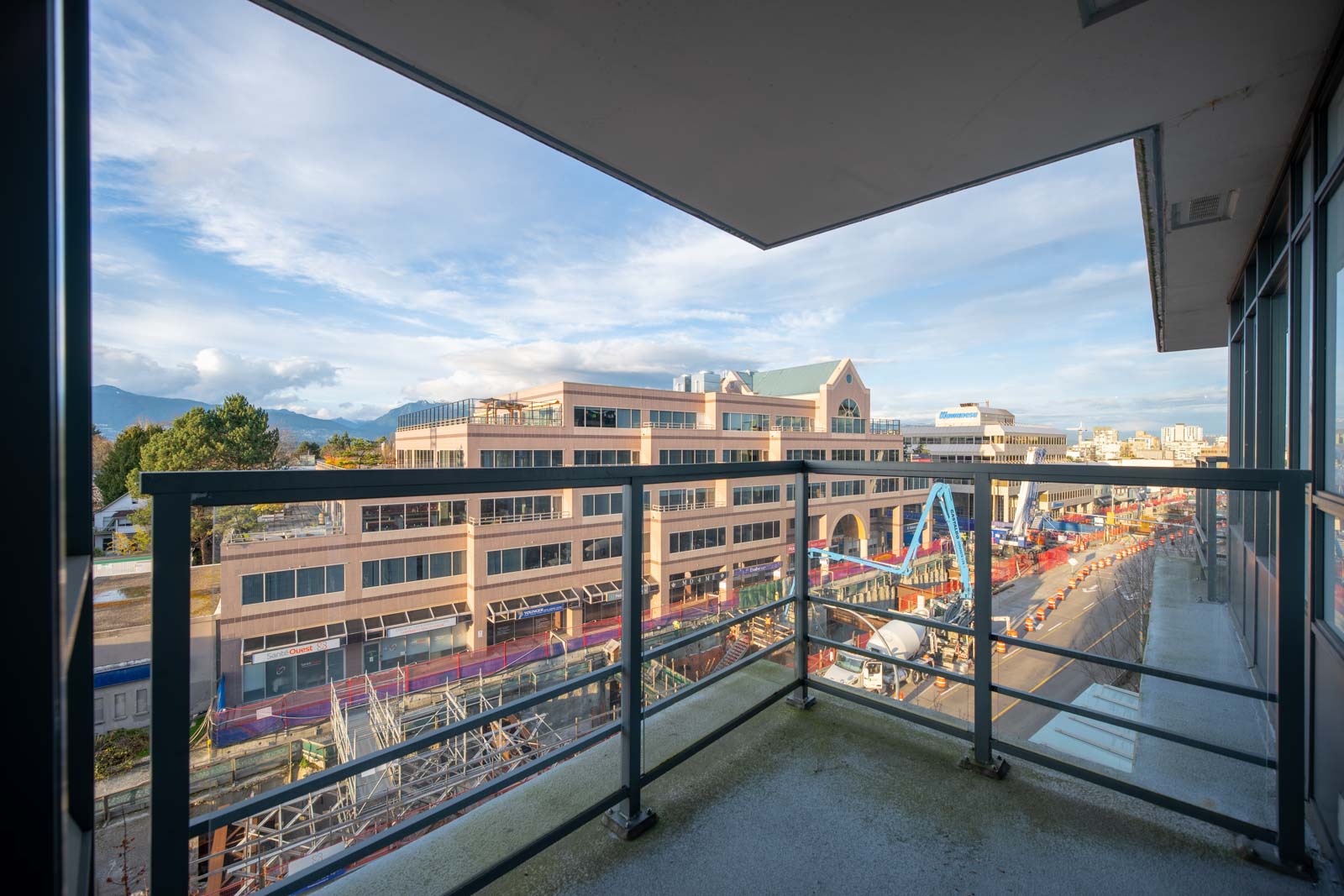 View from a balcony overlooking a construction site on a city street, with a multi-story building and mountains visible in the background under a partly cloudy sky.