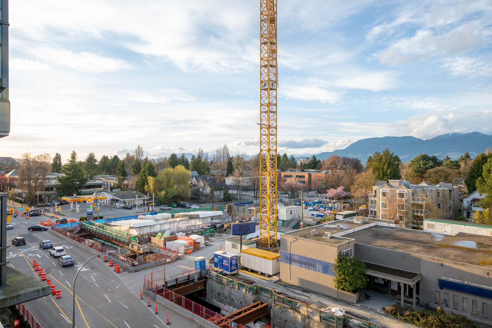A construction site with a large yellow crane, surrounded by buildings, trees, and mountains in the background, next to a road with traffic cones and cars.