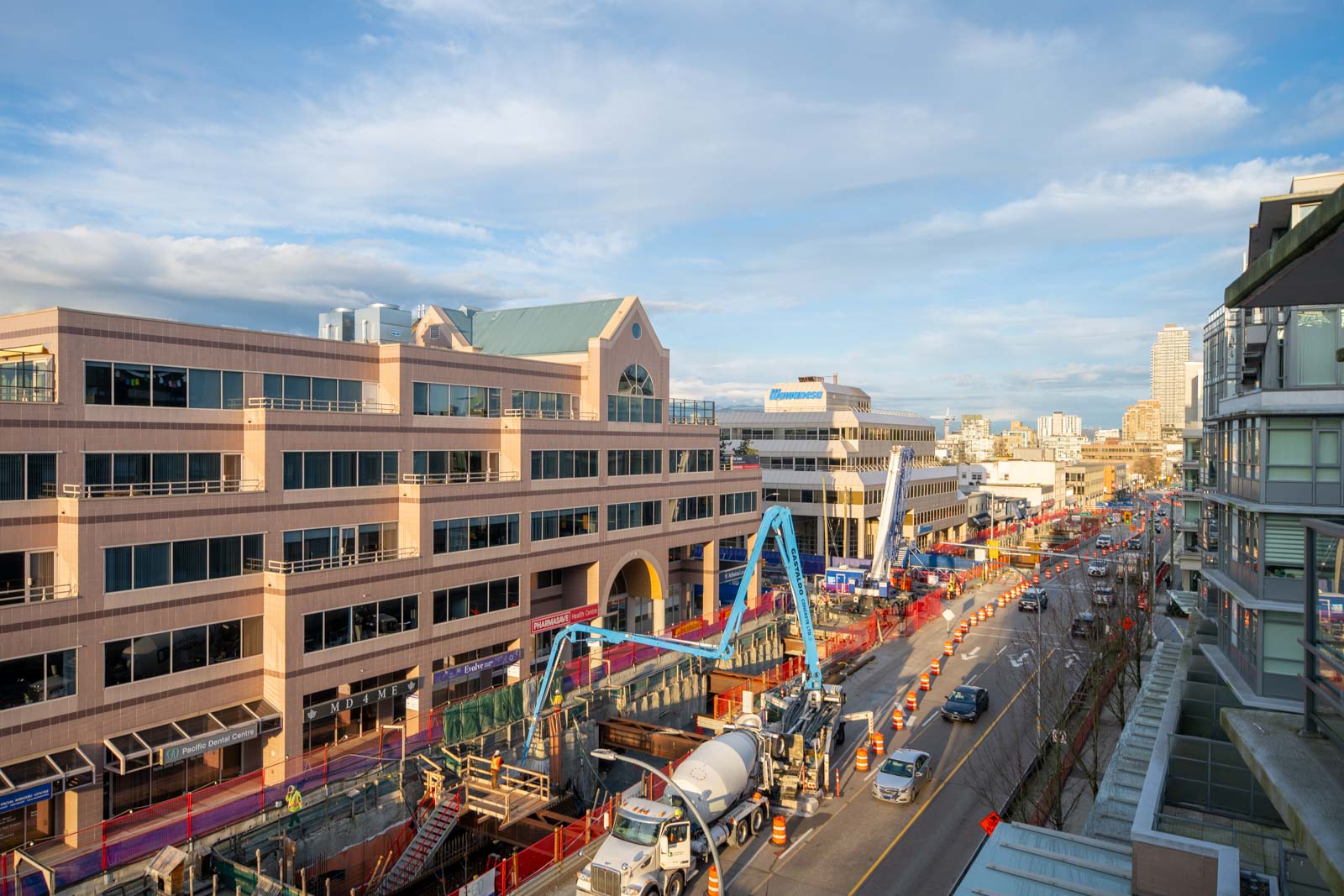 A city street with road construction underway, including cement trucks and equipment, bordered by office and residential buildings under a clear sky.