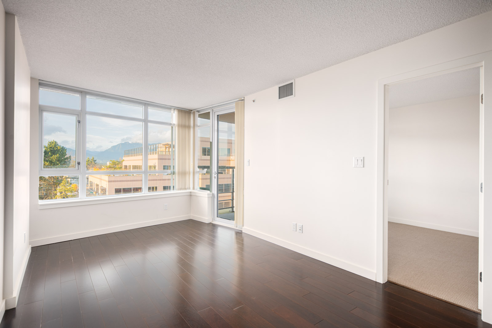 Empty modern apartment room with large windows, dark wood floors, white walls, and an open doorway leading to a carpeted room.
