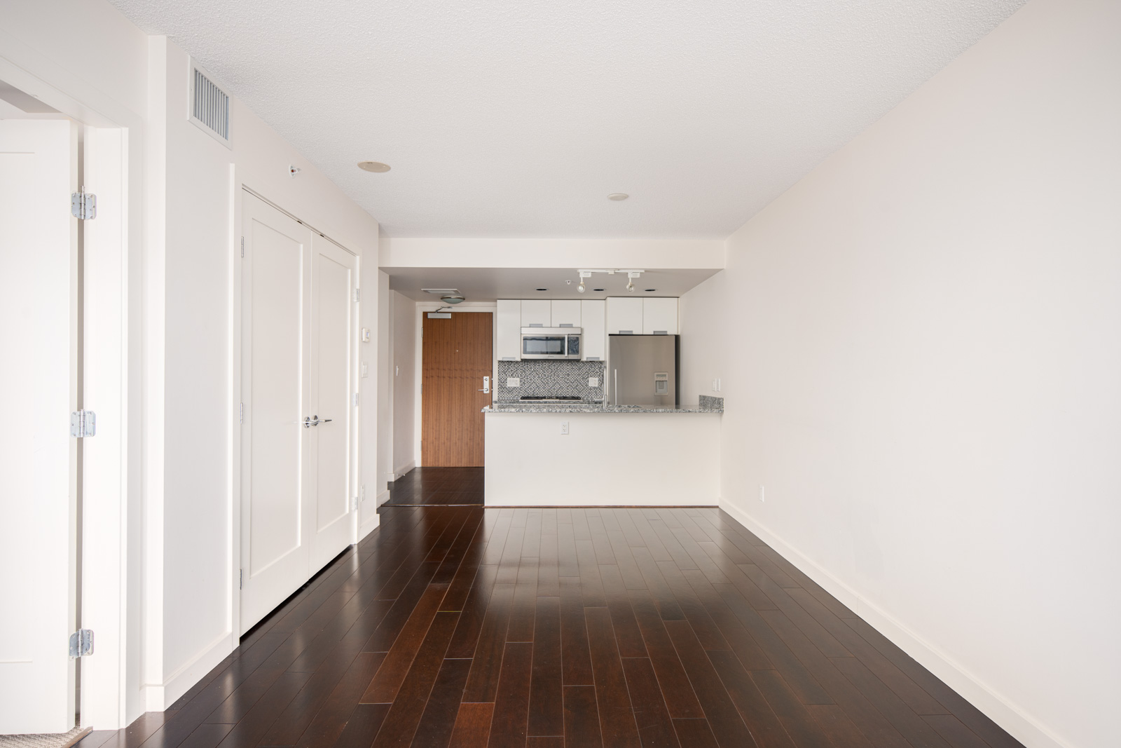 Unfurnished apartment with dark wood floors, white walls, and a modern kitchen featuring stainless steel appliances and granite countertops.