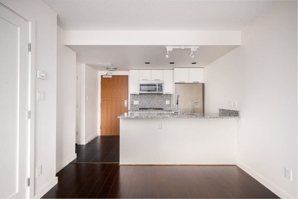 Modern apartment kitchen with white cabinets, stainless steel appliances, granite countertop, and dark wood floors, viewed from the living area with a white wall on the right.