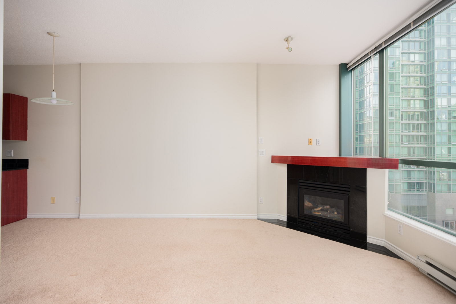 Unfurnished living room with beige carpet, a corner gas fireplace, large floor-to-ceiling windows, and a partial view of an adjacent high-rise building.