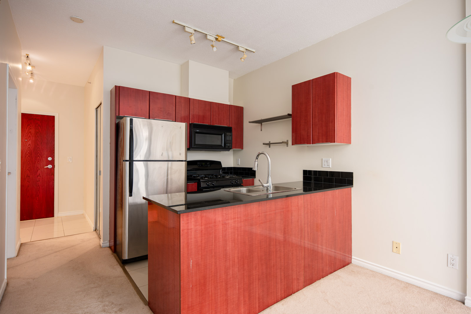 Modern kitchen with red wood cabinets, stainless steel appliances, black countertops, wall shelves, and a chrome faucet. Hallway with a red door is visible in the background.