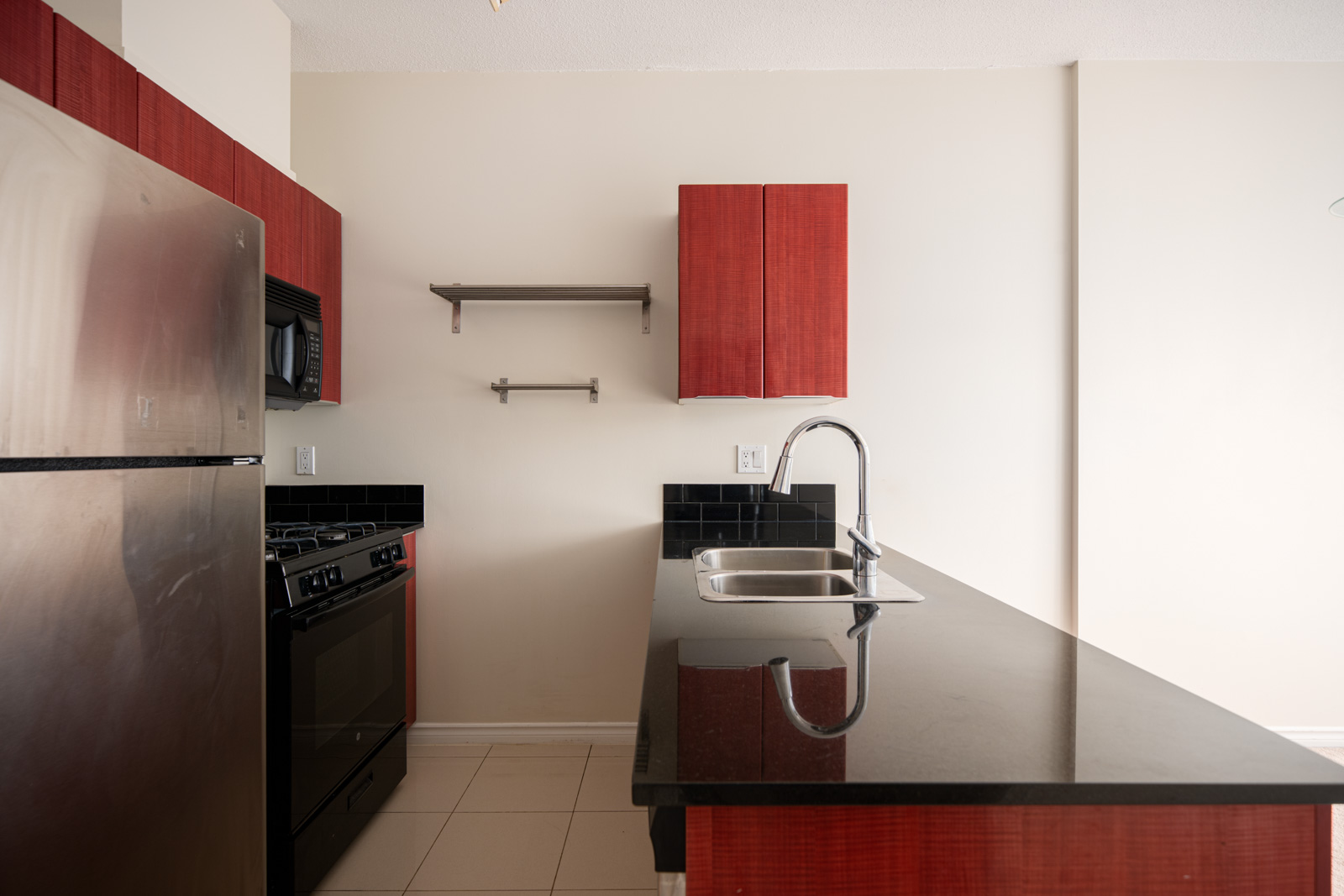 Modern kitchen with stainless steel appliances, black countertop, built-in sink, red cabinets, and open wall shelves against a light-colored wall.