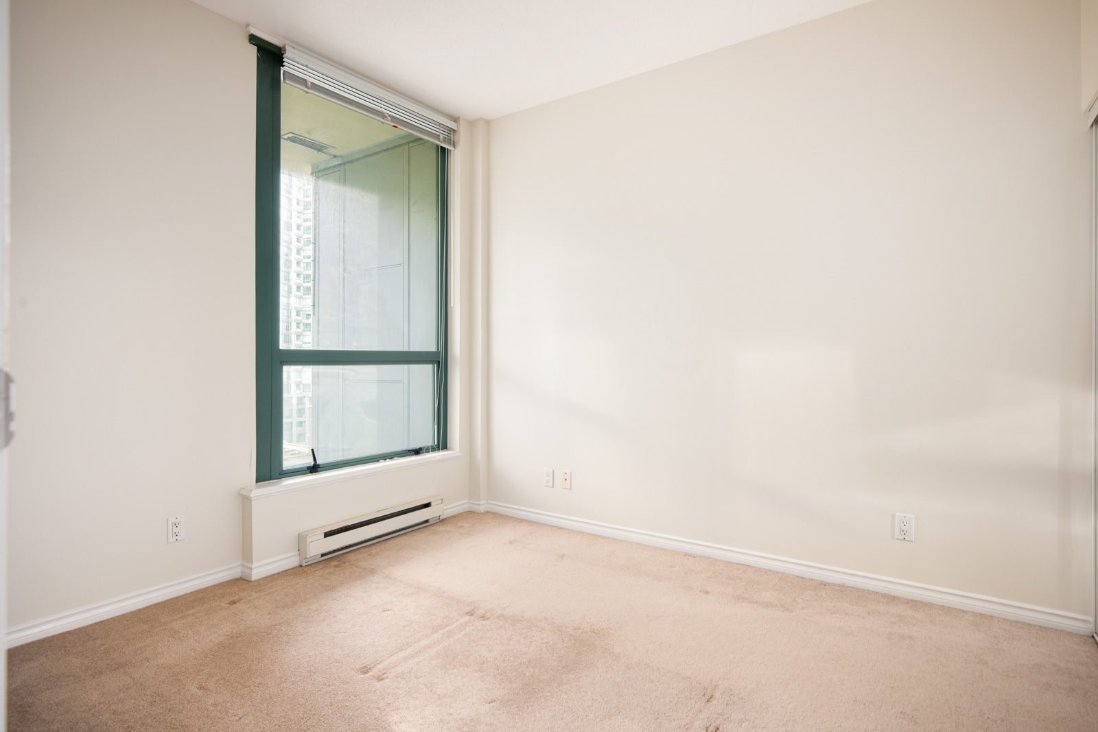 Empty room with beige carpet, large window with vertical blinds, white walls, and baseboard heater below the window.