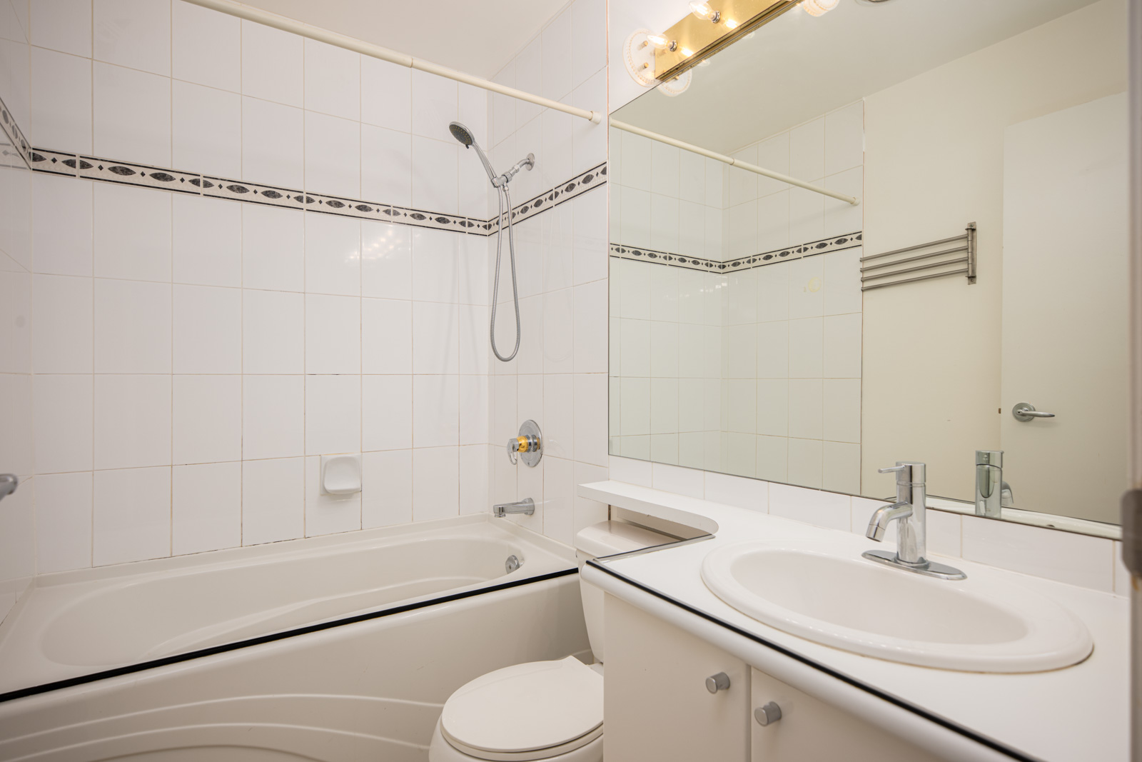 Modern bathroom with white tiles, a bathtub-shower combo, a toilet, a countertop sink, a large mirror, and overhead lighting.