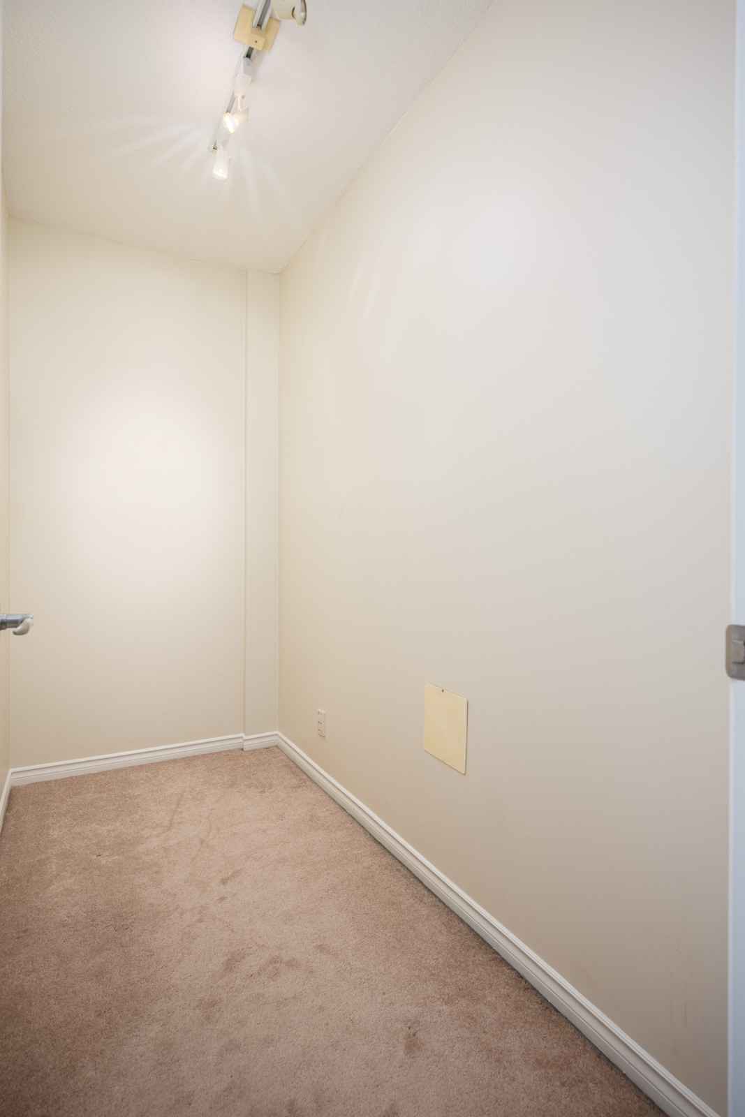 Small, empty room with beige carpet, off-white walls, ceiling track lighting, and a square patch on one wall.