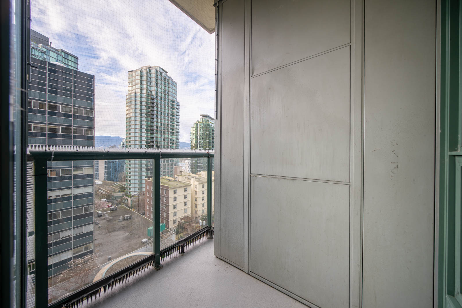 A small balcony with glass railing overlooks city buildings and streets, with tall skyscrapers visible in the background under a partly cloudy sky.