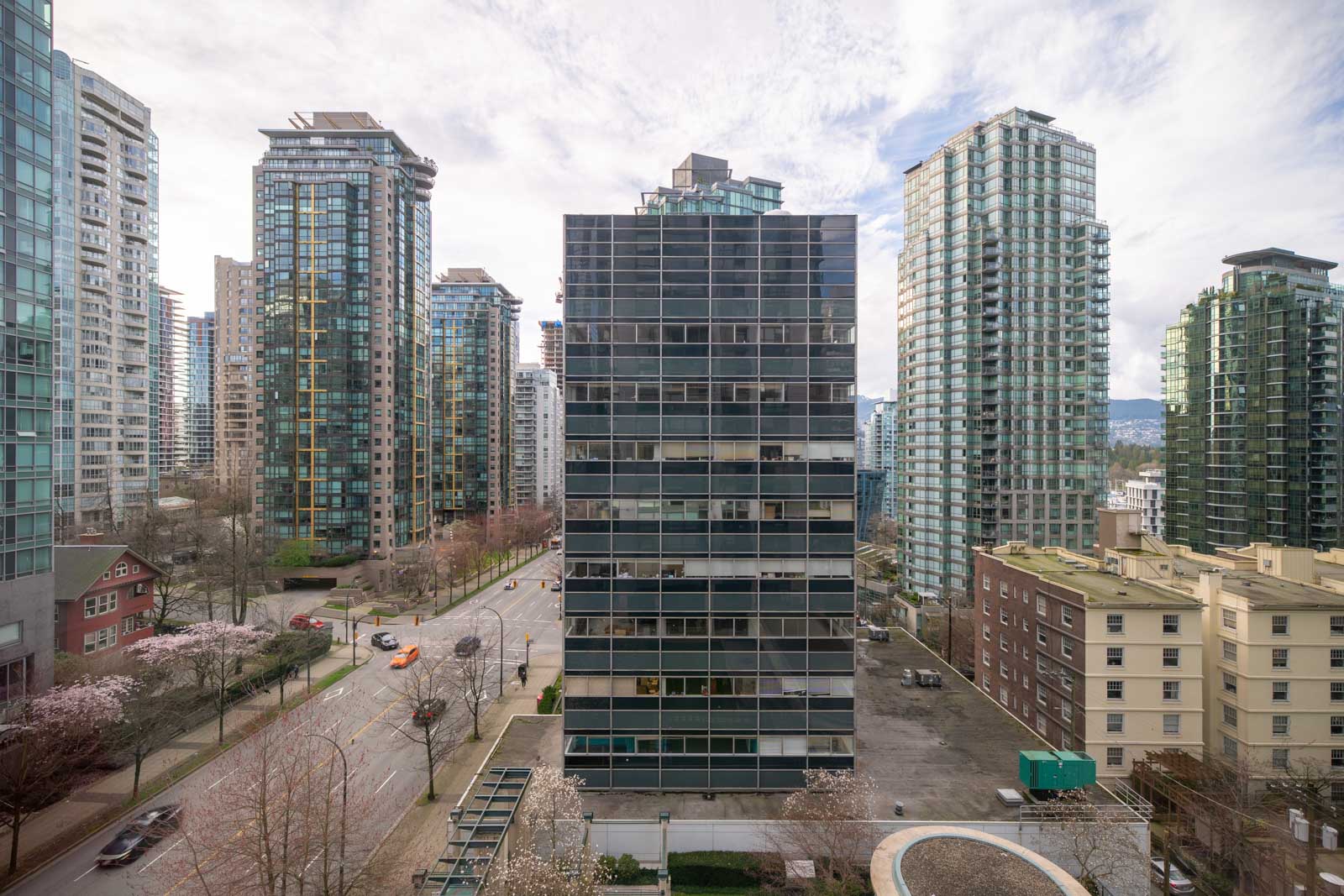 A tall, modern glass office building is centered among high-rise apartment towers in an urban cityscape, with a few cars and trees lining the street below.