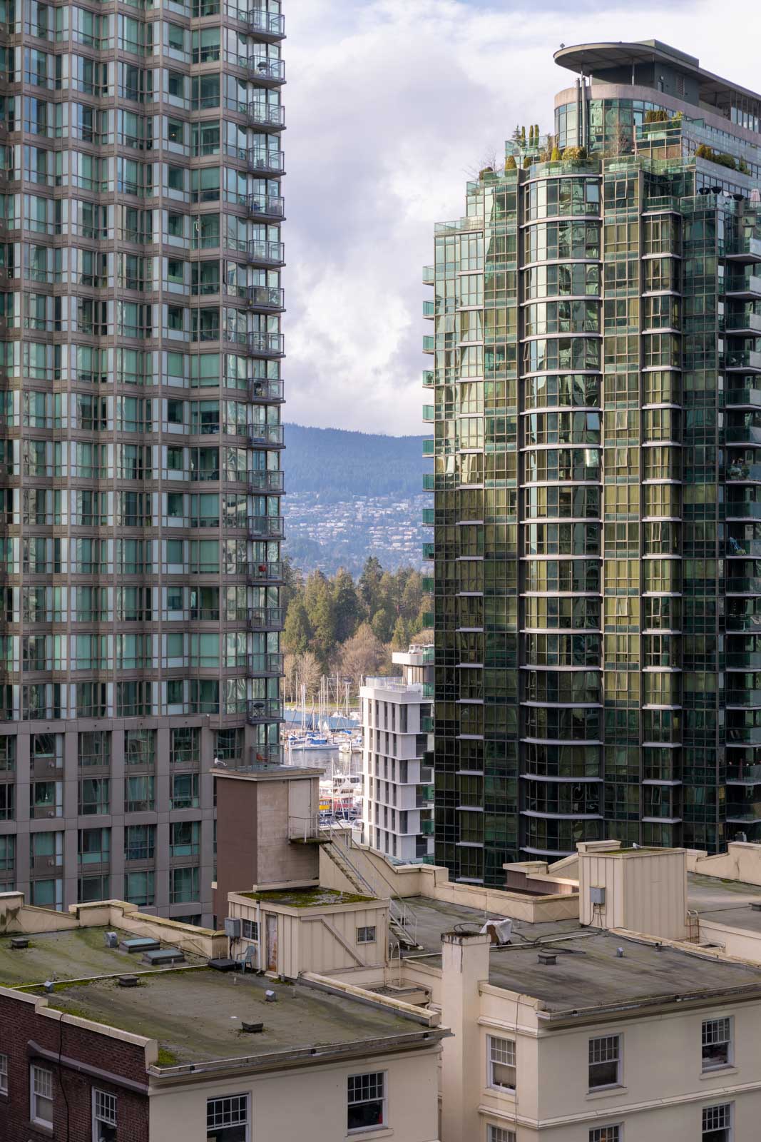 High-rise glass buildings with trees and mountains in the background, viewed above the rooftops of lower, older structures on a cloudy day.