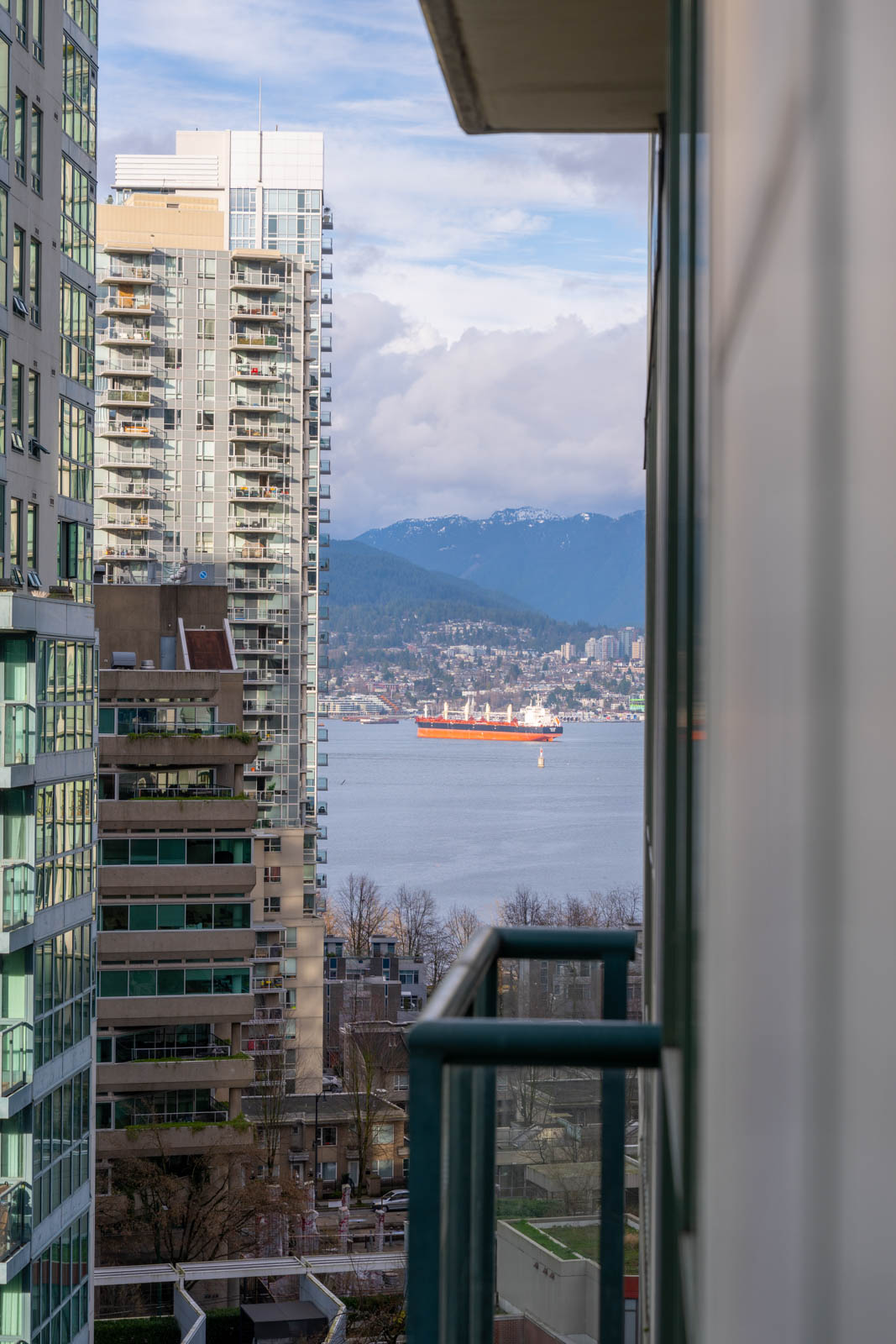 A view between apartment buildings of a large cargo ship on the water, with mountains and cityscape visible in the background under a partly cloudy sky.