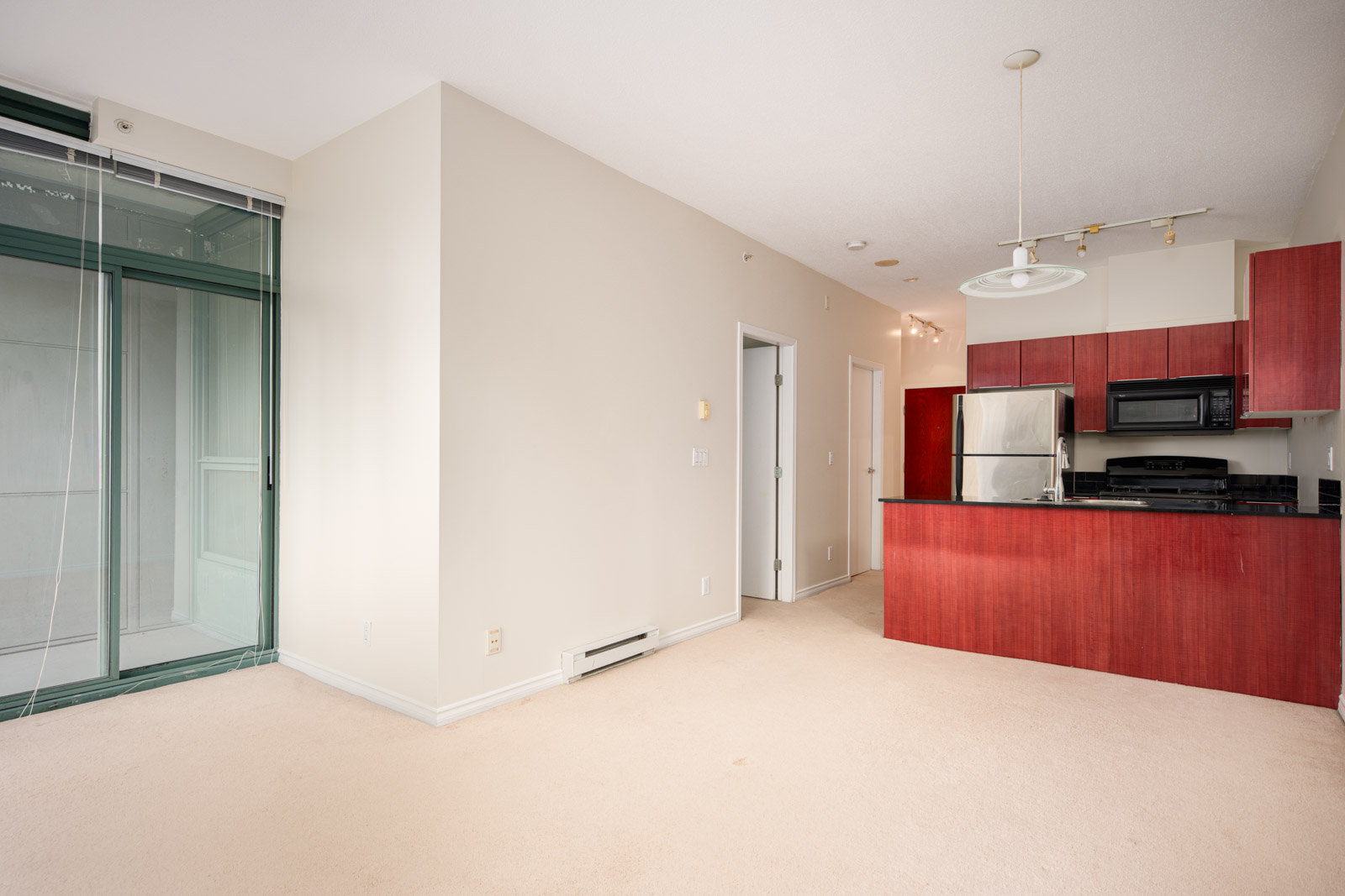 Empty apartment interior with beige carpet, white walls, glass door to balcony, and open kitchen featuring red cabinets and black appliances.