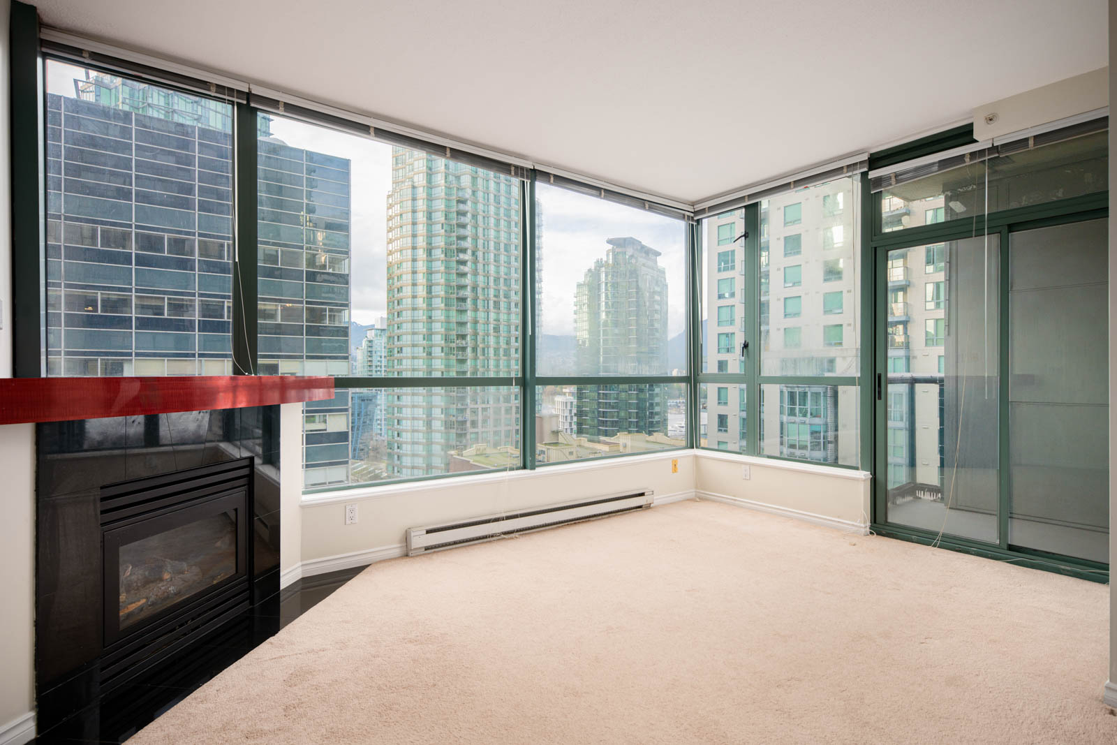A bright corner apartment room with large floor-to-ceiling windows, city views, a glass door to a balcony, beige carpet, and a fireplace with a red mantle.
