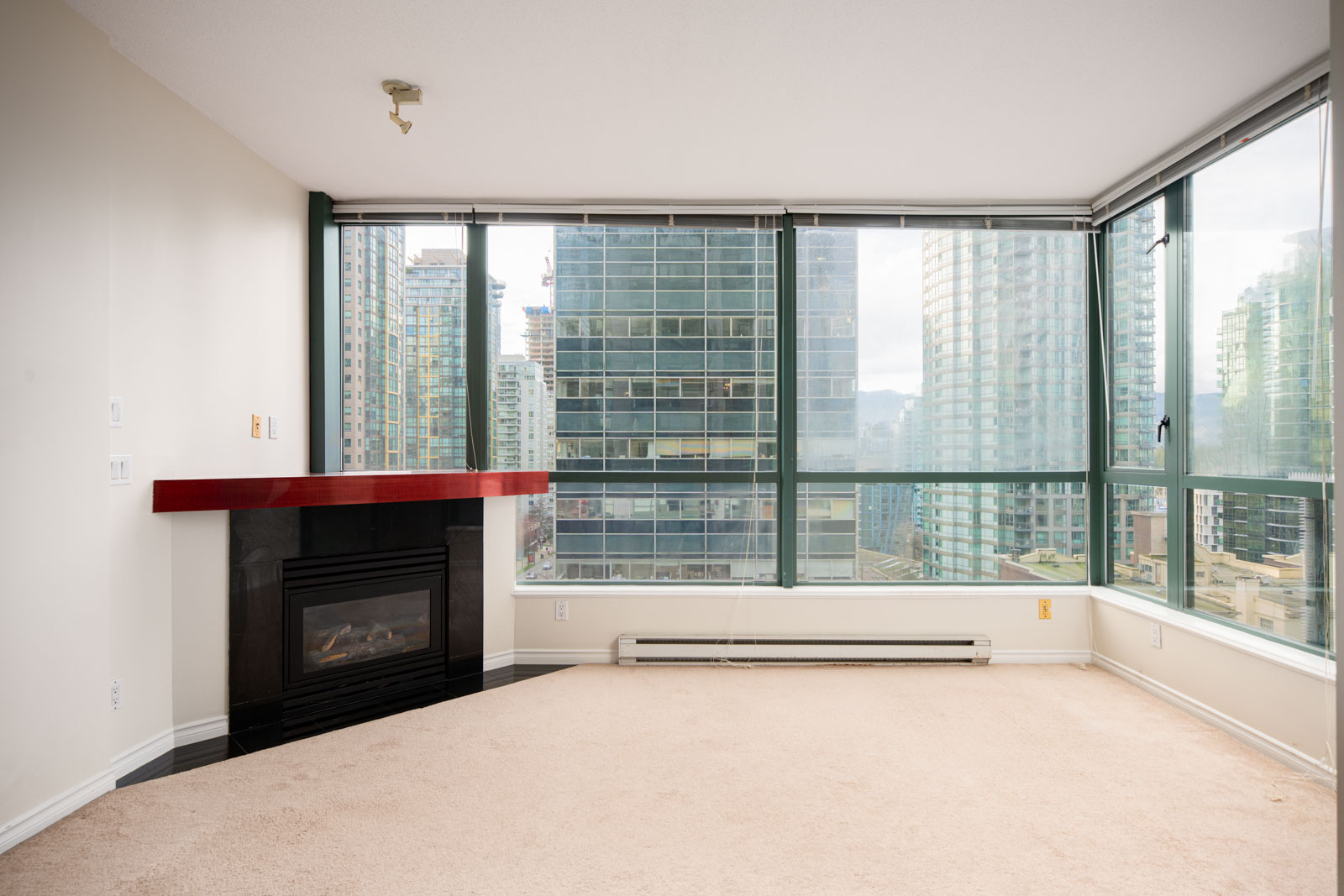 Unfurnished corner apartment with large windows, beige carpet, fireplace with red mantel, and city high-rise views.