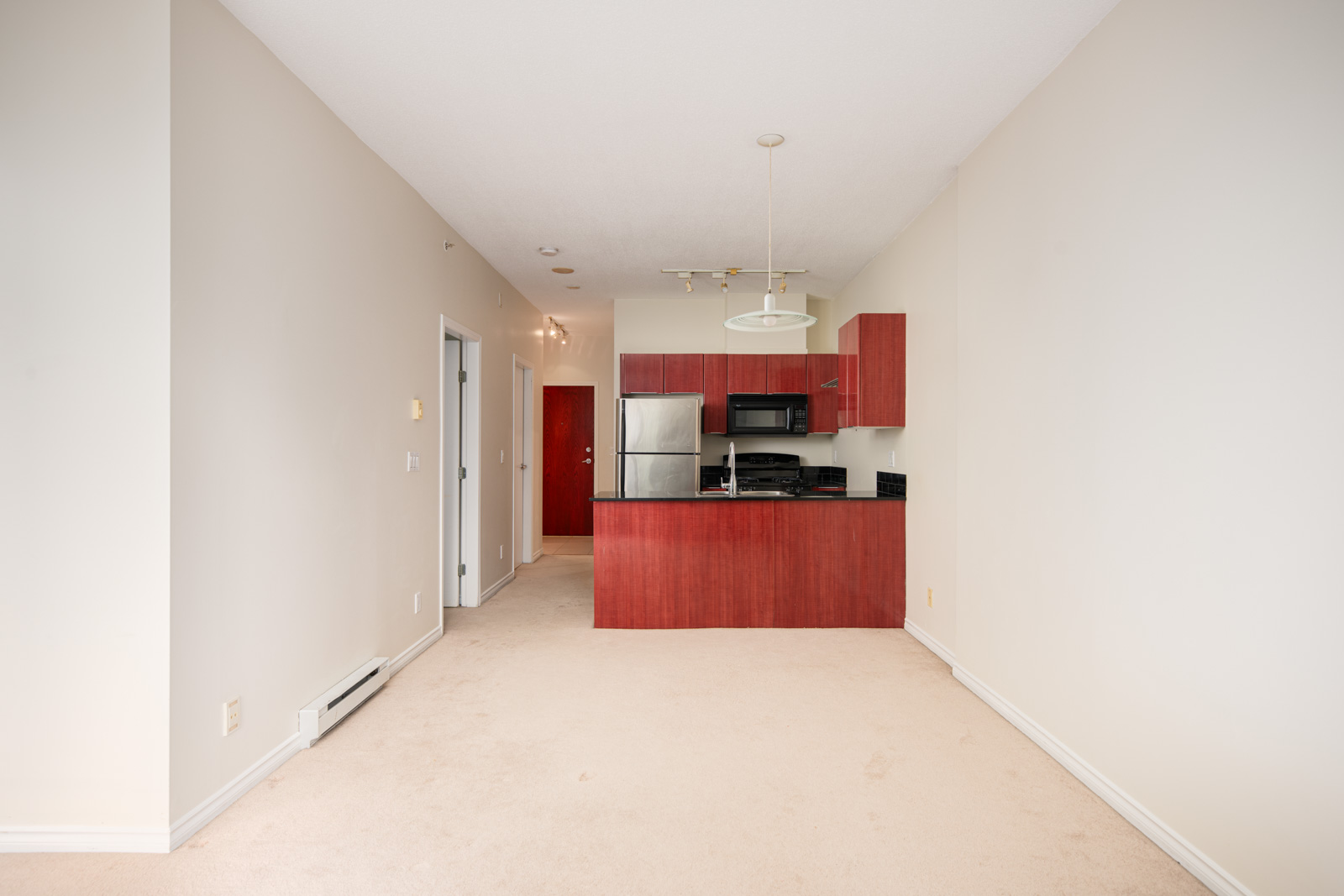 A small apartment kitchen with red cabinets, stainless steel appliances, and beige carpeted flooring, viewed from an empty living area.