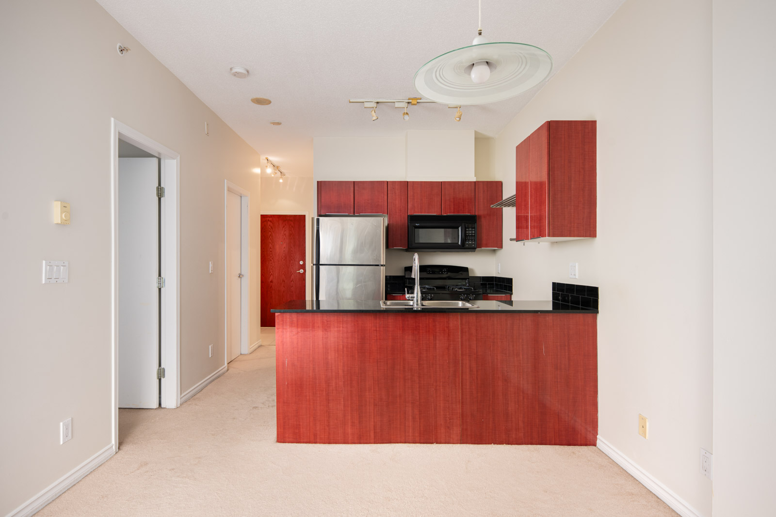 Modern apartment kitchen with red wood cabinets, black countertops, stainless steel appliances, and beige walls, viewed from the living area.