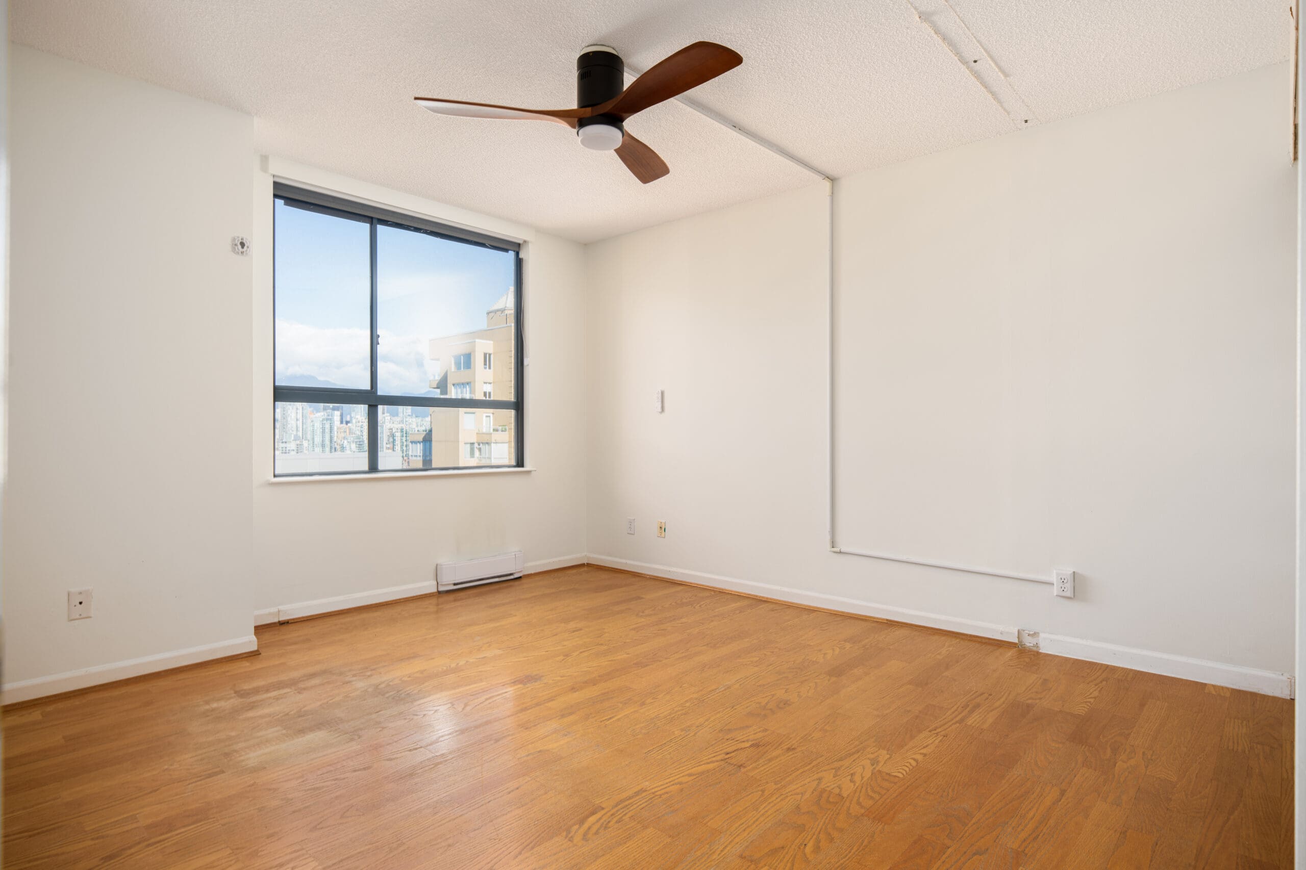 Empty room with white walls, wood floor, ceiling fan, and large window showing a city view. Natural light fills the space.
