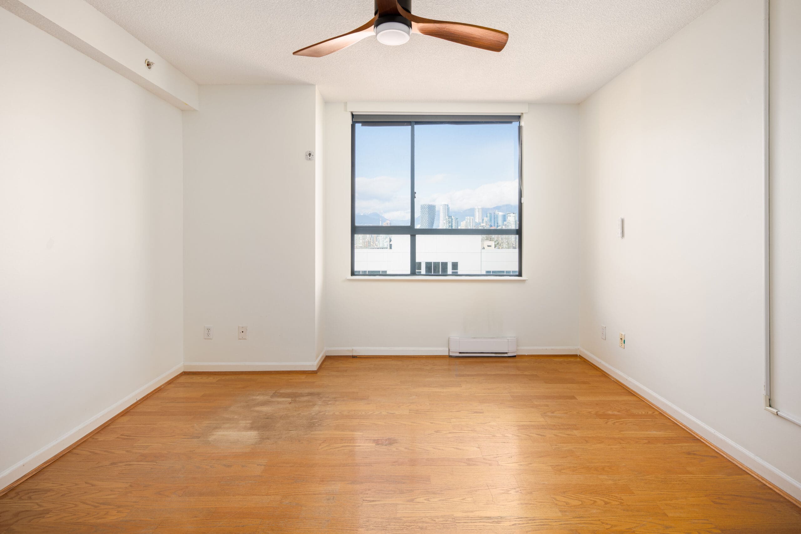Empty room with hardwood floors, a ceiling fan, white walls, and a large window with a city view in the background.