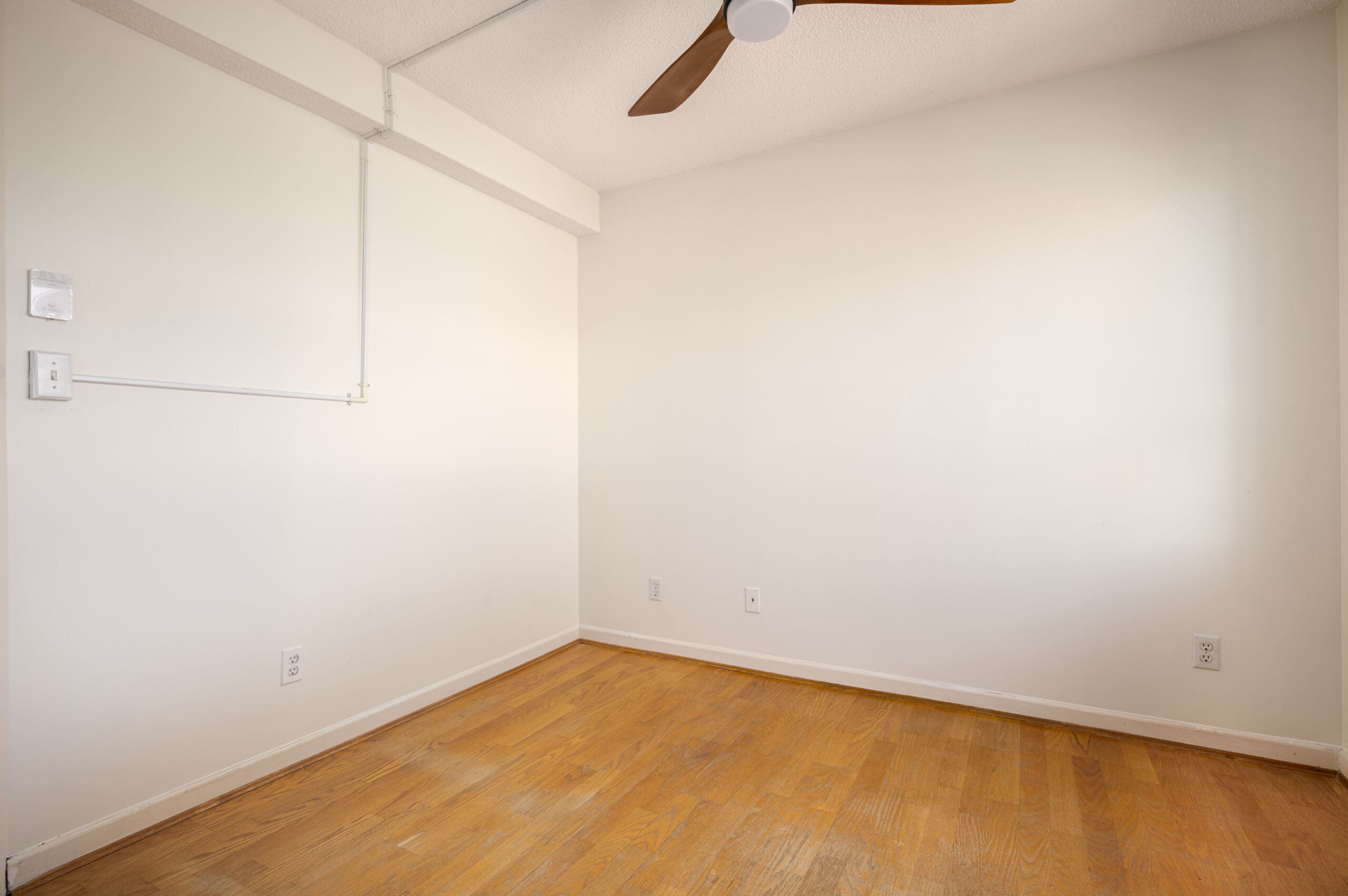 Empty room with hardwood floor, white walls, several electrical outlets, and a ceiling fan with wooden blades.