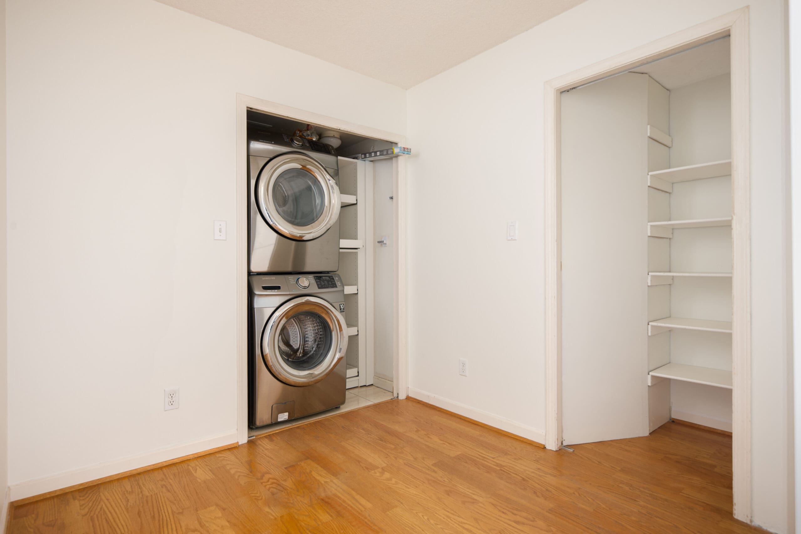 A laundry area with a stacked washer and dryer next to an open closet with built-in shelves, both in a room with hardwood flooring and white walls.