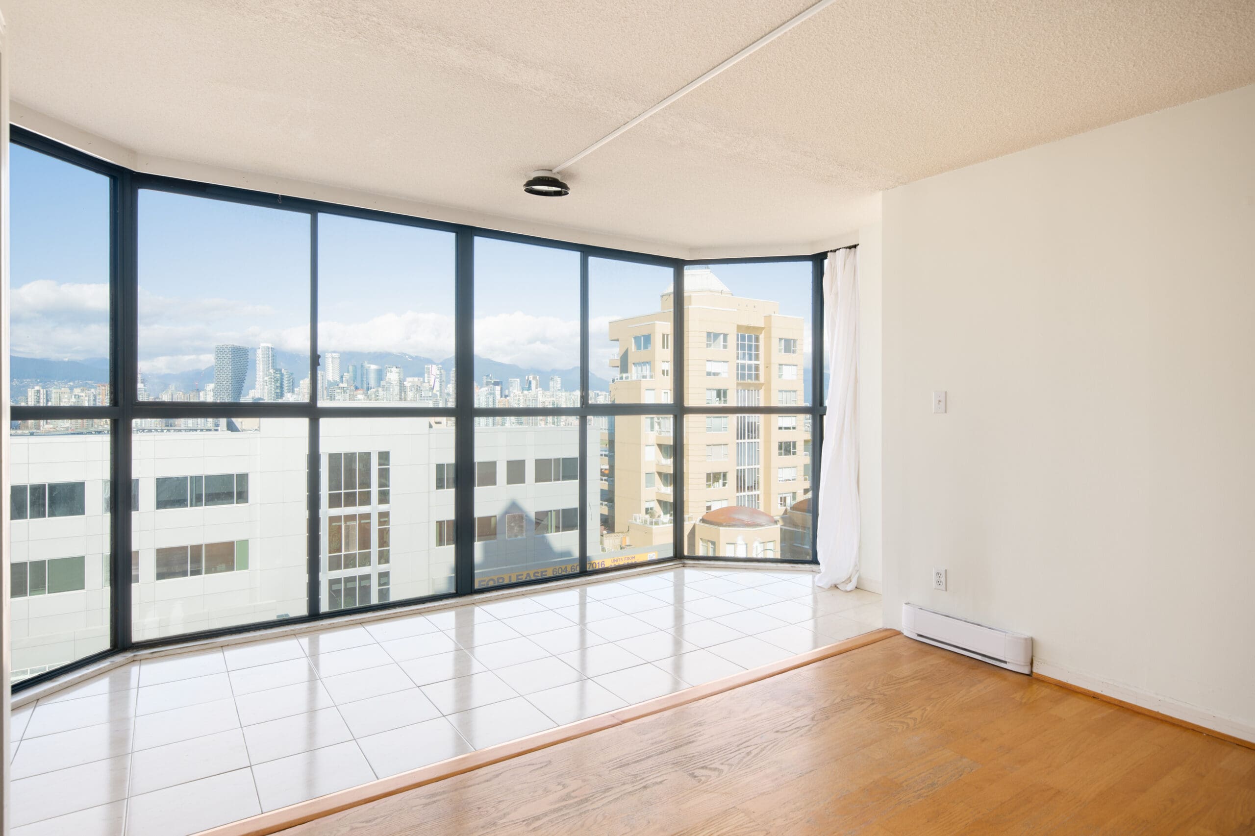 Corner room with large floor-to-ceiling windows, tiled and hardwood flooring, and a view of city buildings and mountains in the distance.
