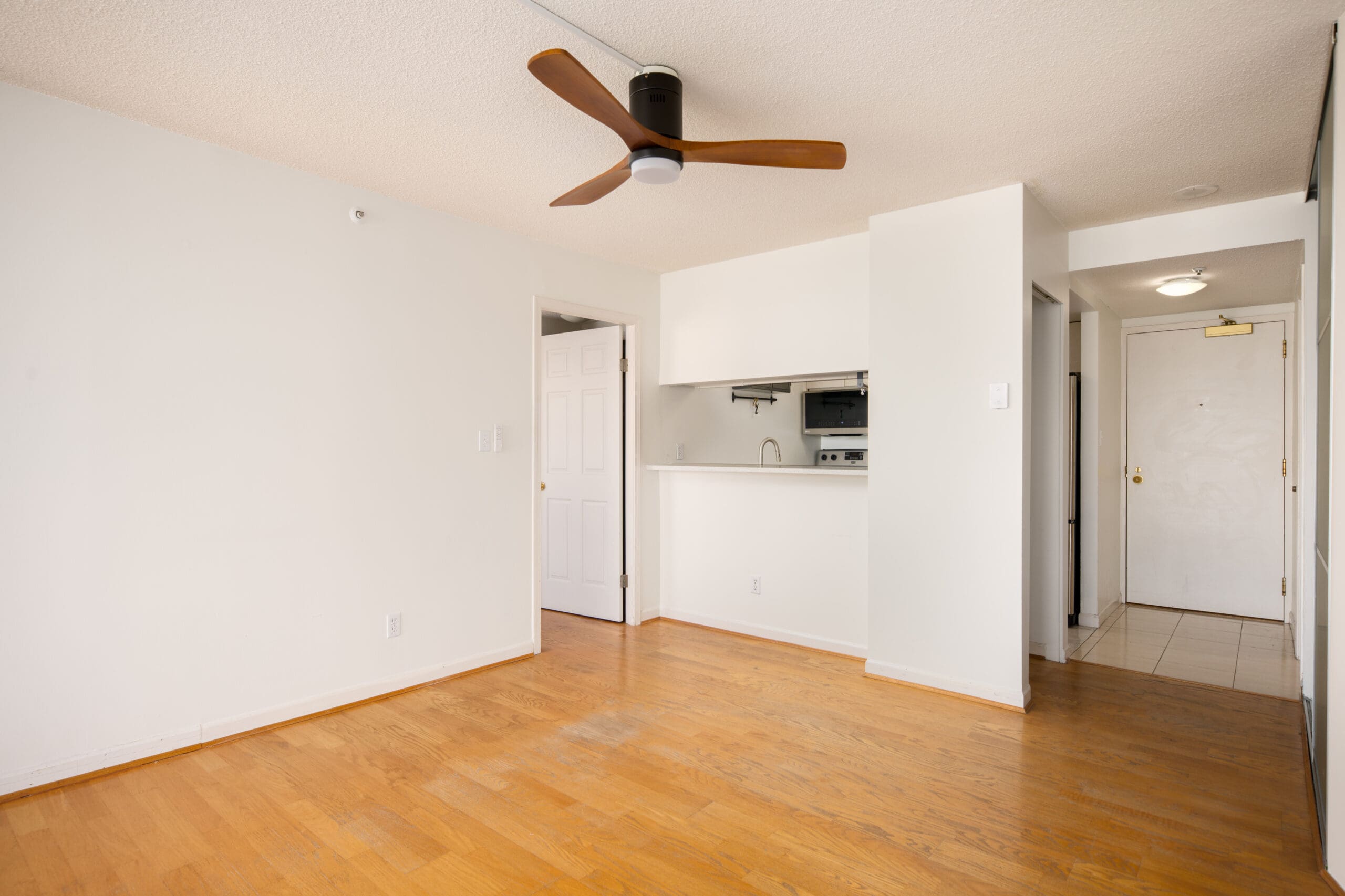 Unfurnished apartment with light wood flooring, white walls, a ceiling fan, small kitchen with pass-through window, and a hallway leading to the front door.
