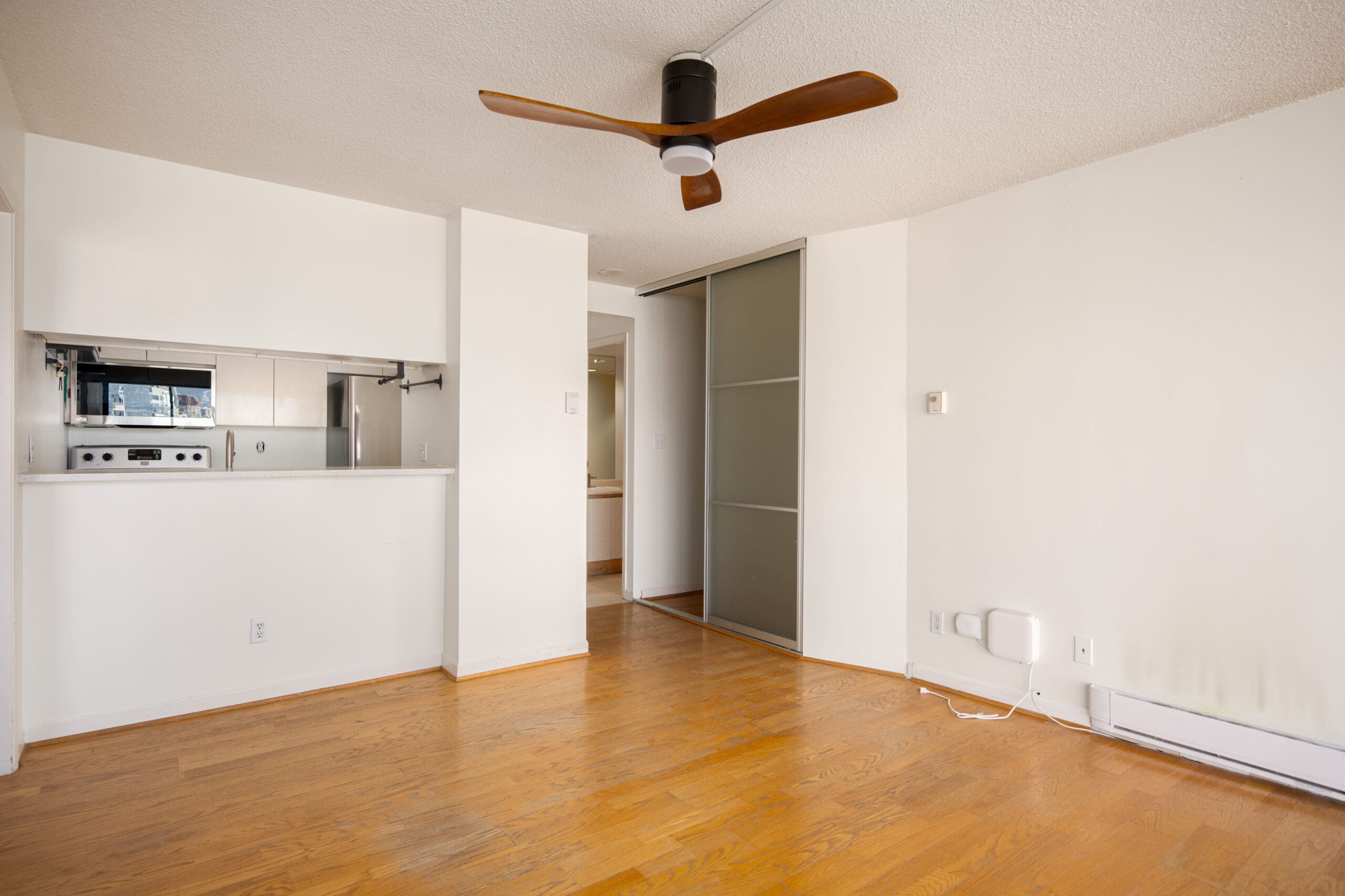 Unfurnished room with hardwood floors, white walls, ceiling fan, sliding closet doors, and a partial view of the kitchen area.