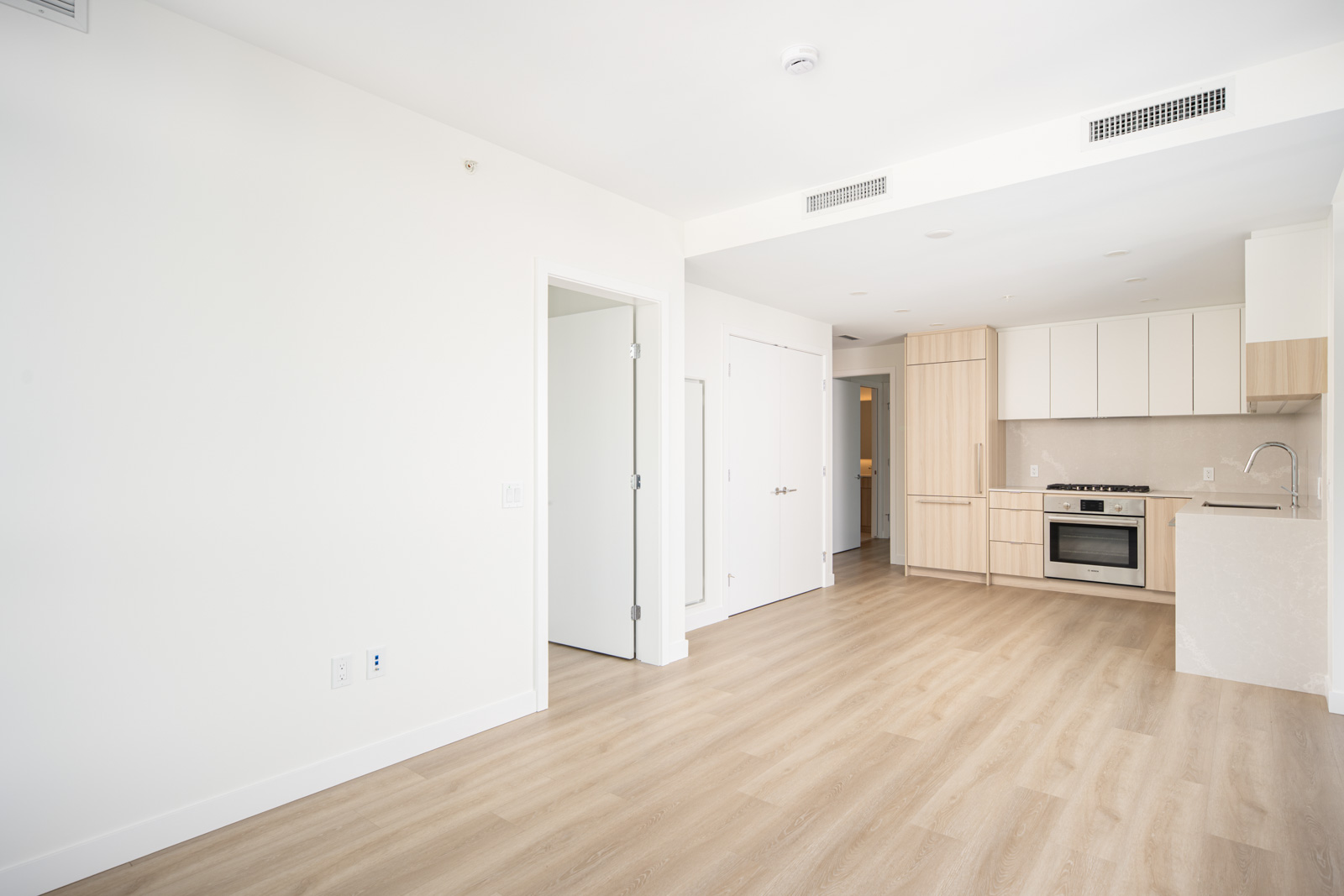 A modern, unfurnished apartment with light wood flooring, white walls, and an open kitchen featuring built-in appliances and light wood cabinets.