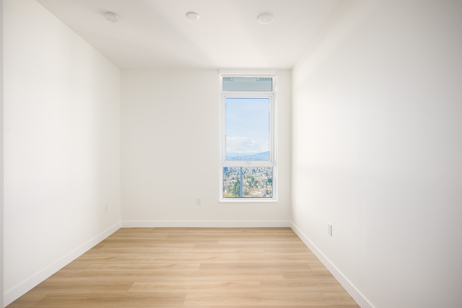 Empty white room with light wood flooring and a single window showing a cityscape and mountains under a blue sky.