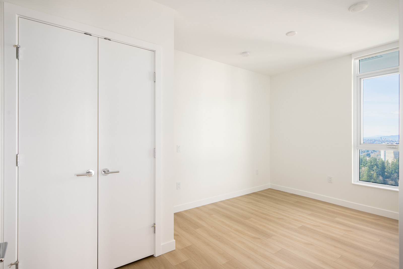 Empty, unfurnished room with light wood floors, white walls, double doors, and a large window letting in natural light with a partial city and tree view.