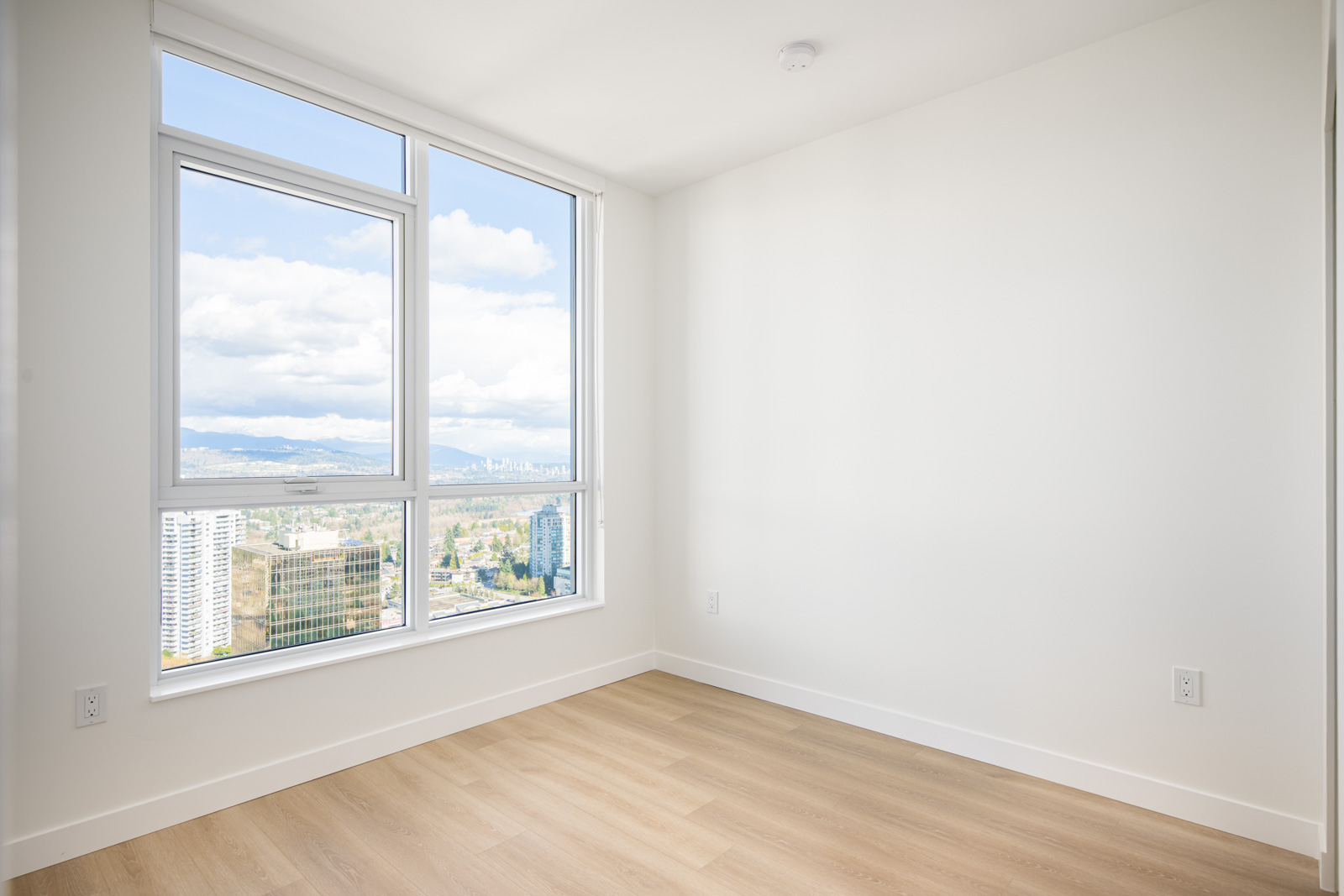 Empty room with white walls, light wood floor, and large window showing a cityscape and cloudy sky outside.