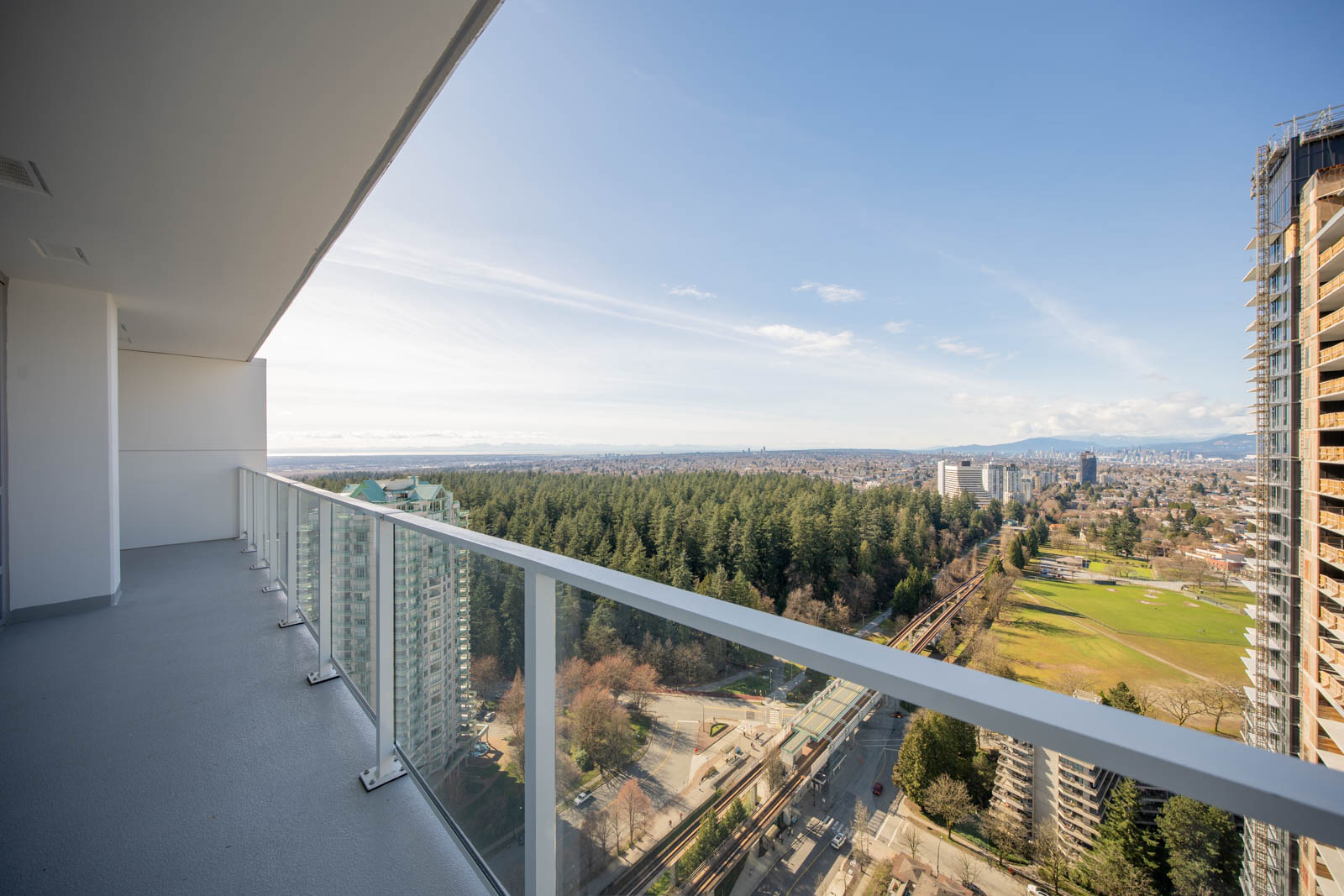 High-rise balcony with glass railing overlooking a cityscape, green park, forest area, and distant mountains under a clear sky.