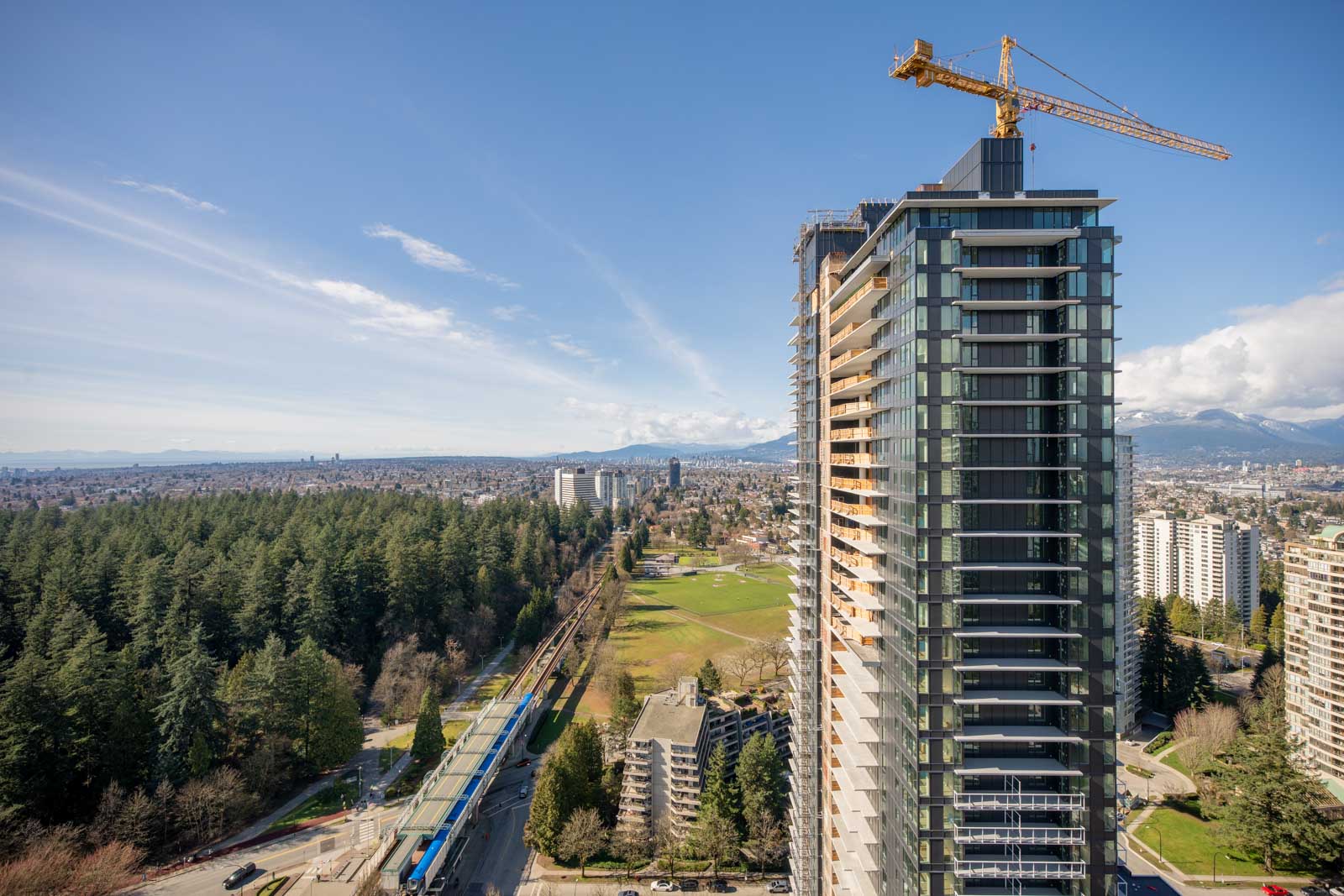 A tall modern high-rise with a construction crane is seen near a park, surrounded by trees and other buildings, with mountains visible in the background under a partly cloudy sky.