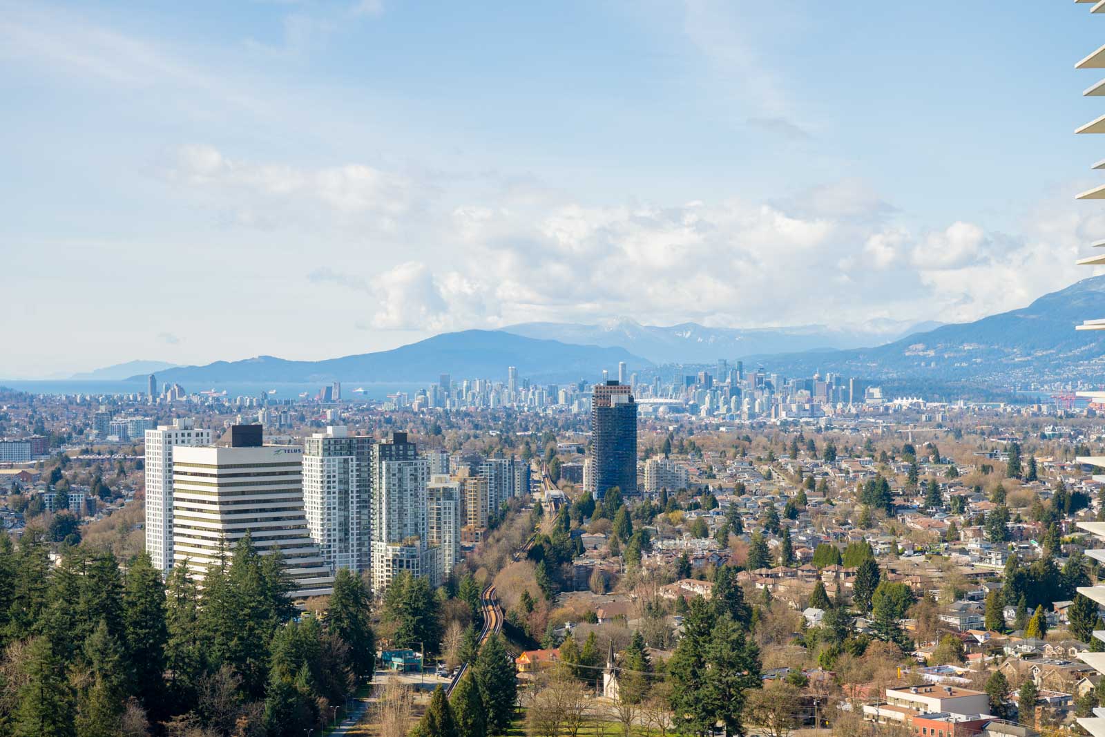 A wide view of a cityscape with tall buildings, dense residential areas, trees, and mountains in the background under a partly cloudy sky.