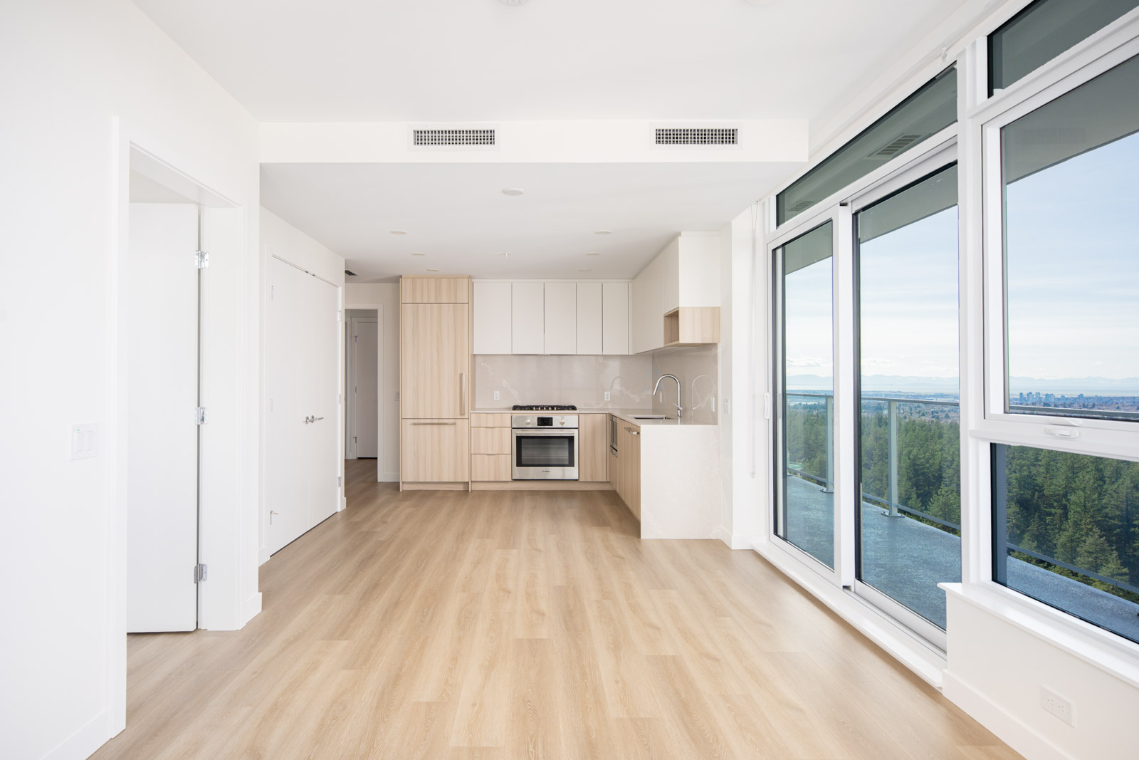 Modern, unfurnished apartment with light wood floors, a kitchen with built-in appliances, white cabinets, and large windows offering a view of trees and a distant city skyline.