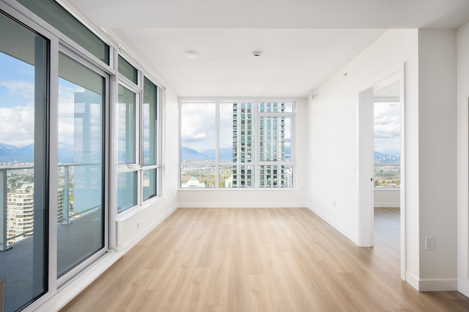 Bright, empty apartment room with large floor-to-ceiling windows, light wood flooring, and a city and mountain view. Adjacent room partially visible through an open doorway.