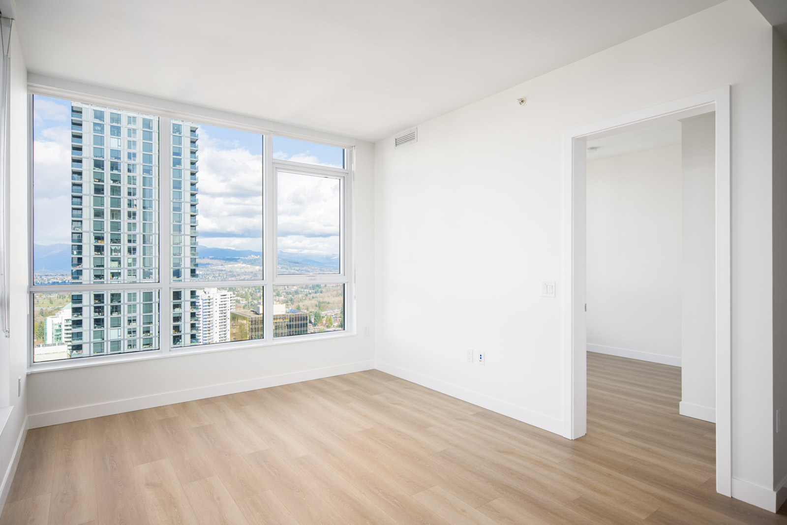 Empty modern apartment room with large windows, light wood flooring, white walls, and a view of city buildings and mountains outside.