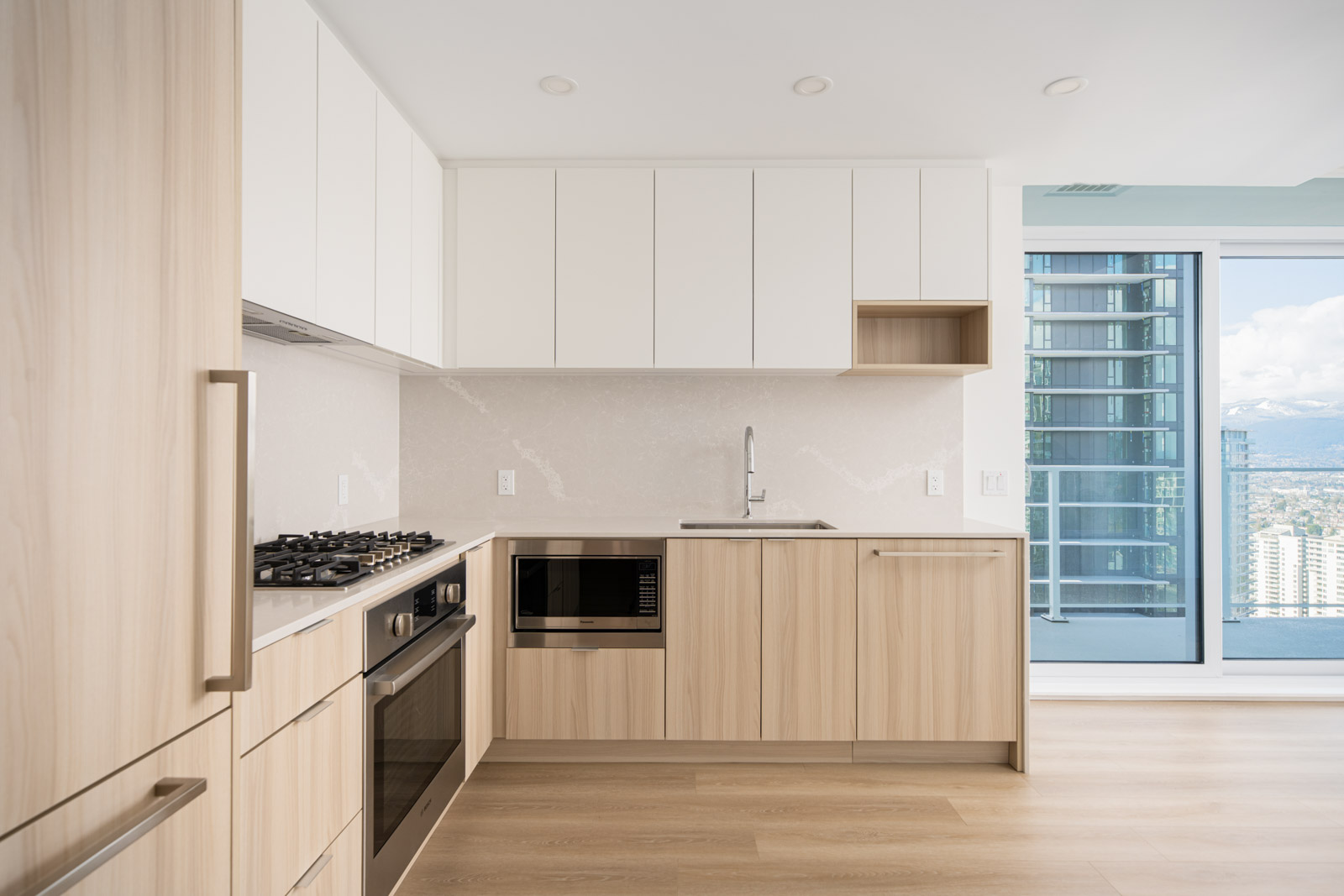 Modern kitchen with light wood cabinetry, built-in oven, gas cooktop, microwave, and white upper cabinets. Large window and balcony with city and mountain views in the background.