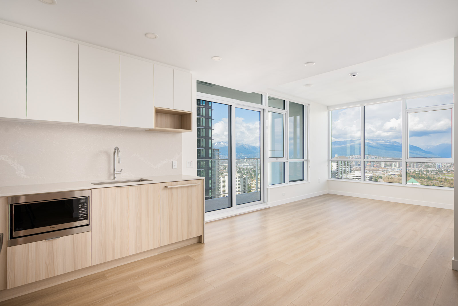 Modern, empty apartment with light wood floors, built-in kitchen cabinets, microwave, large windows, and a balcony overlooking a cityscape with mountains in the background.