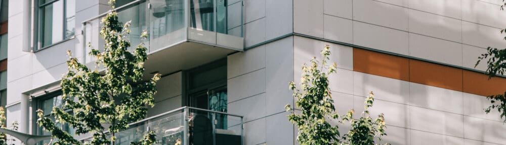 Modern apartment building exterior with glass balconies and horizontal orange accent stripes, partially obscured by green leafy trees in the foreground.