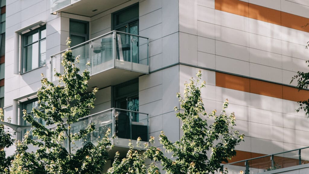 Modern apartment building exterior with glass balconies and horizontal orange accent stripes, partially obscured by green leafy trees in the foreground.
