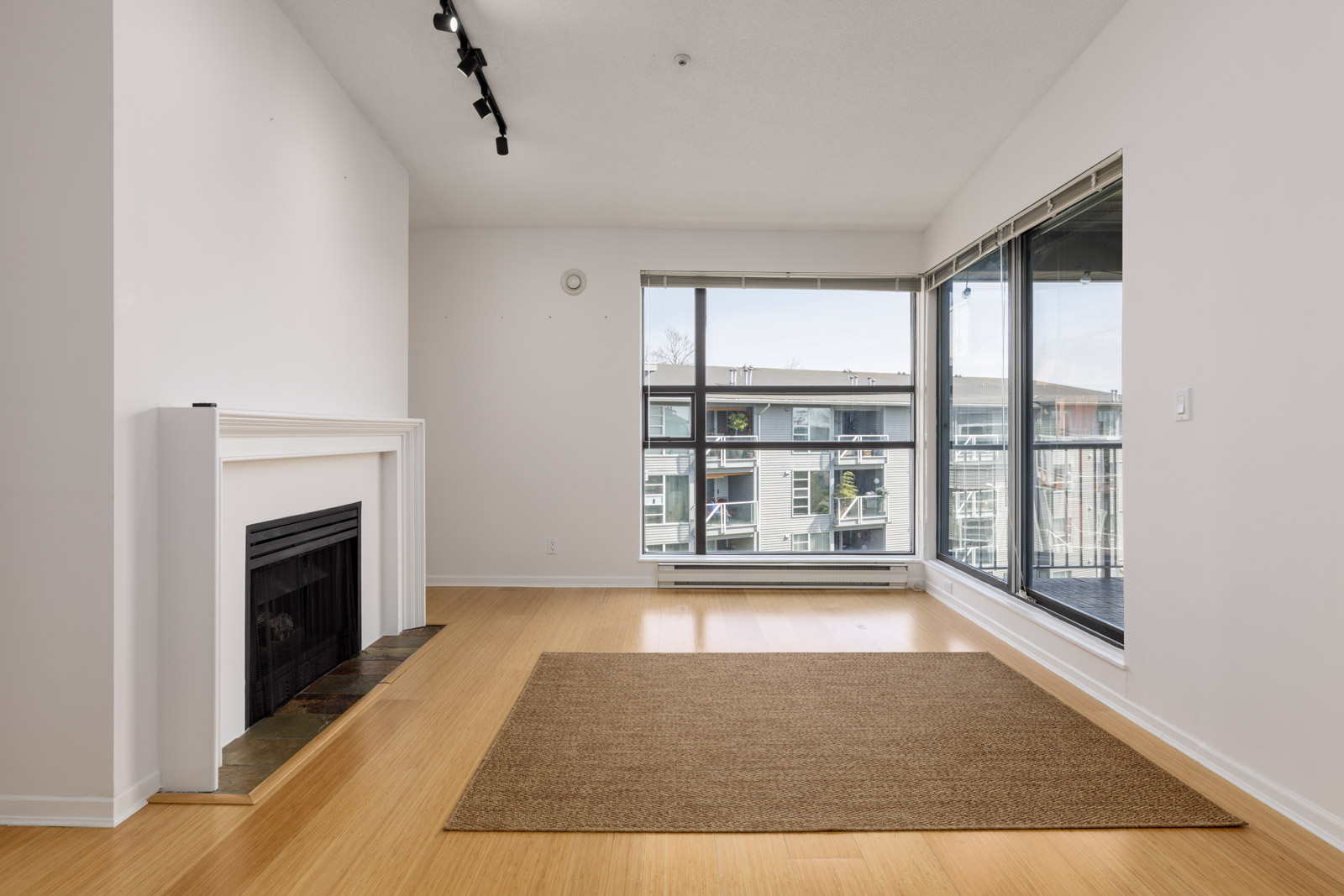 Empty living room with a fireplace, wood flooring, a brown area rug, and large windows overlooking a modern apartment building.