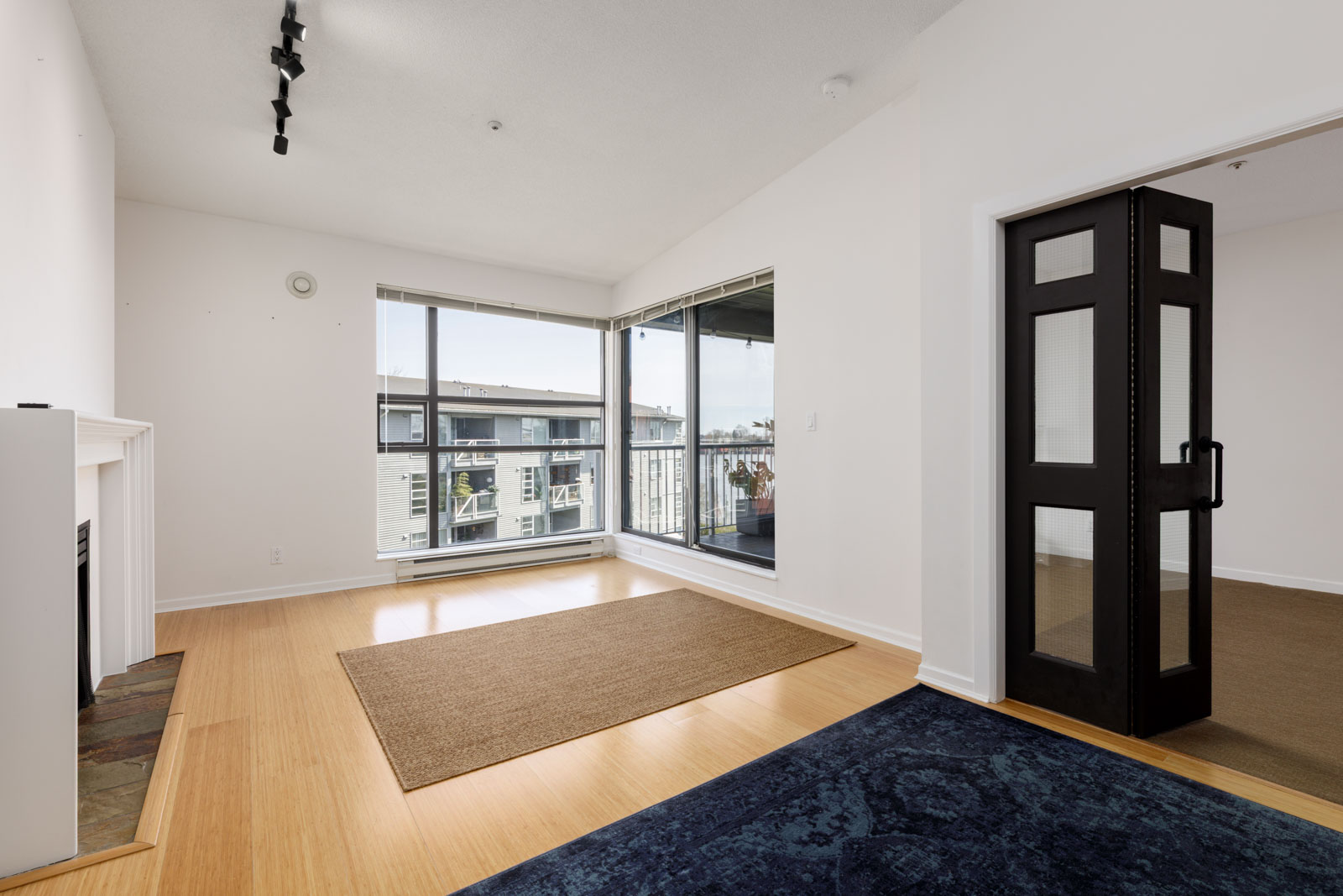 Bright, empty living room with light wood floors, a fireplace, large windows, a brown rug, and glass doors leading to another room and a balcony.