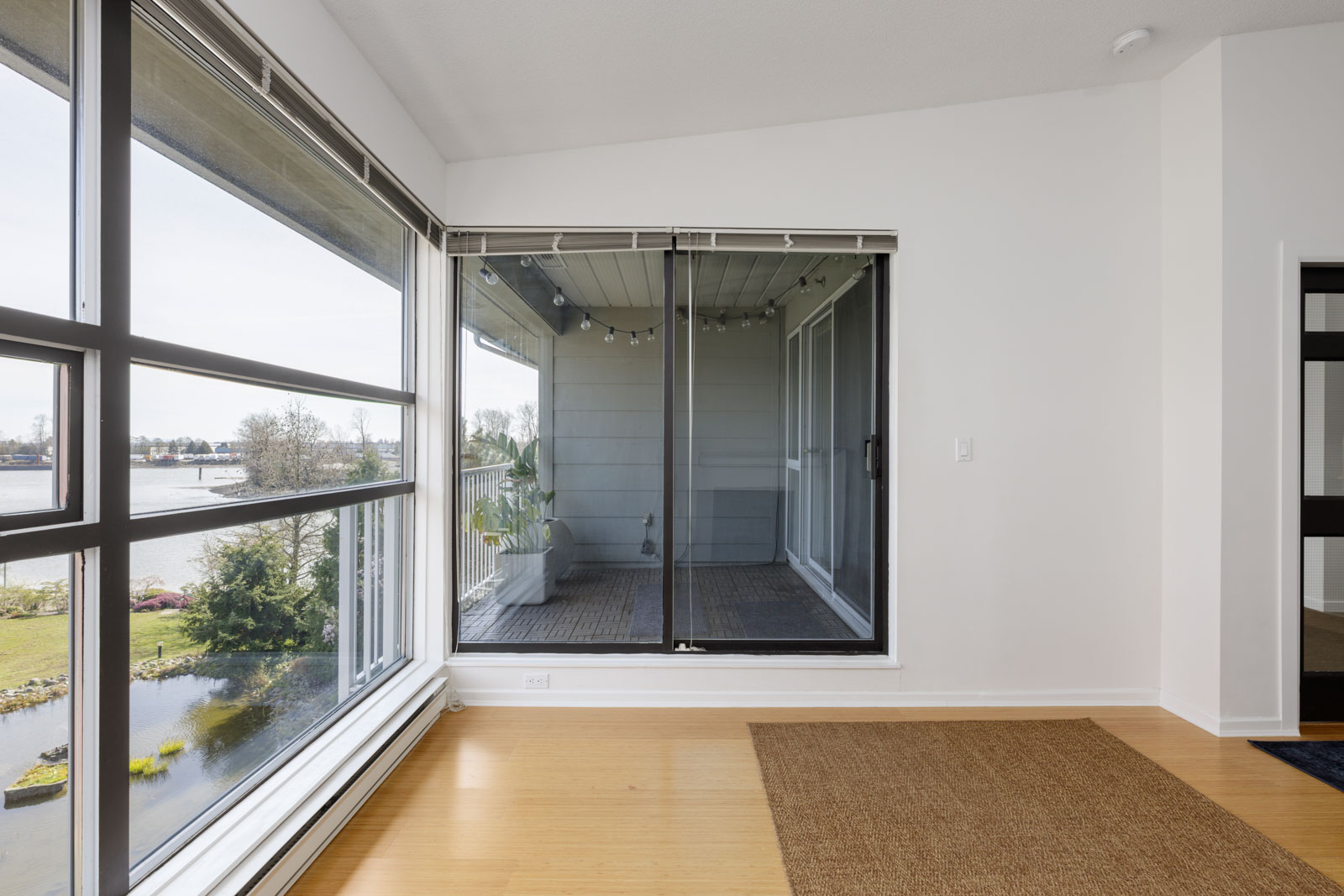 A room with large windows and a sliding glass door leading to a covered balcony, overlooking a body of water and greenery outside.
