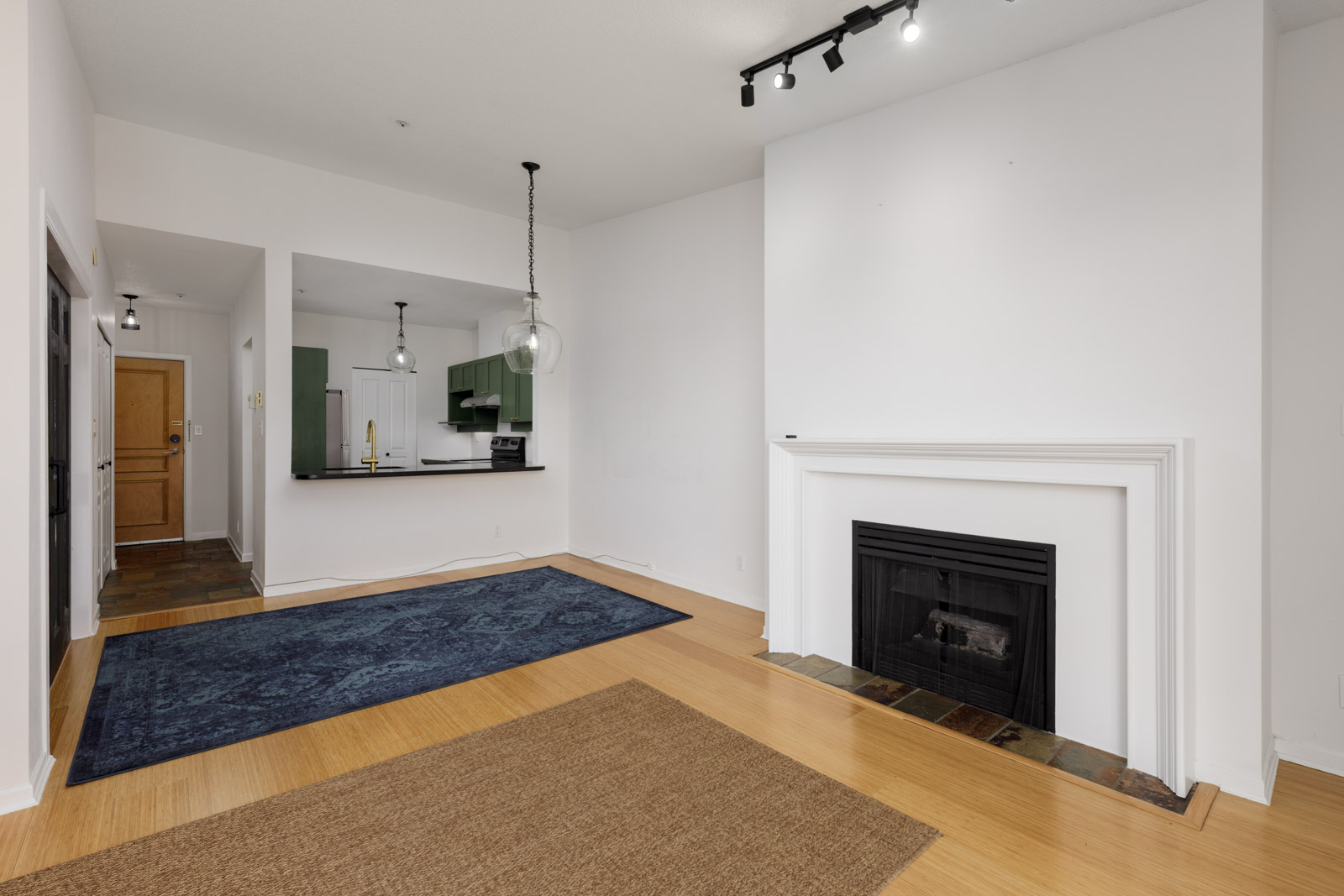 Modern living room with light wood flooring, white walls, a black fireplace, two rugs, and an open view of a kitchen with green cabinets and pendant lighting.